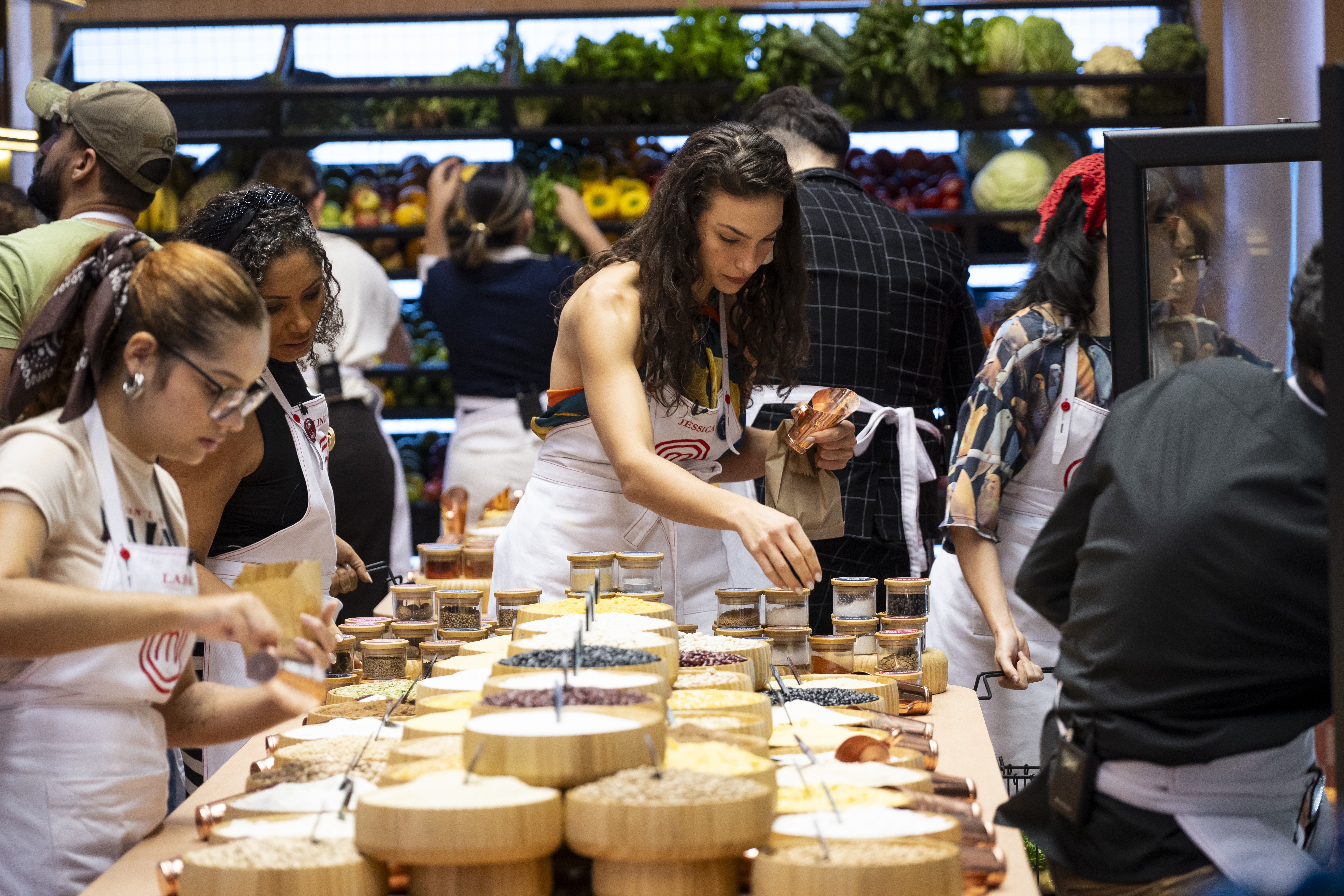 Mulheres com aventais brancos preparam ingredientes em uma bancada cheia de potes e tigelas com grãos e temperos. Ao fundo, outras pessoas e prateleiras com frutas e vegetais frescos em um mercado ou supermercado