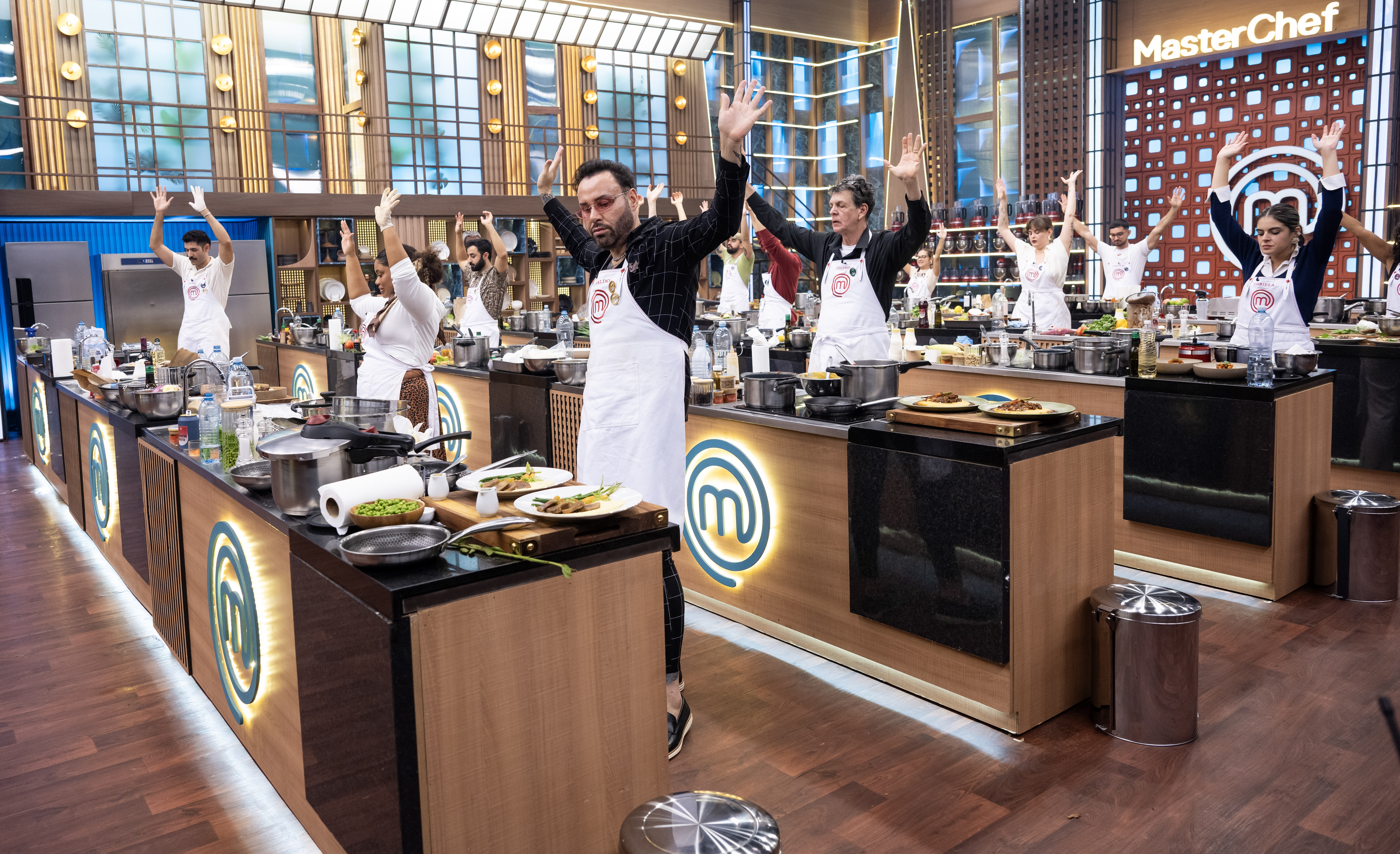 Competidores do MasterChef, usando aventais brancos, levantam os braços em comemoração ou instrução, em uma cozinha profissional com bancadas de madeira e metal, panelas e pratos prontos. Ao fundo, o logo do programa.