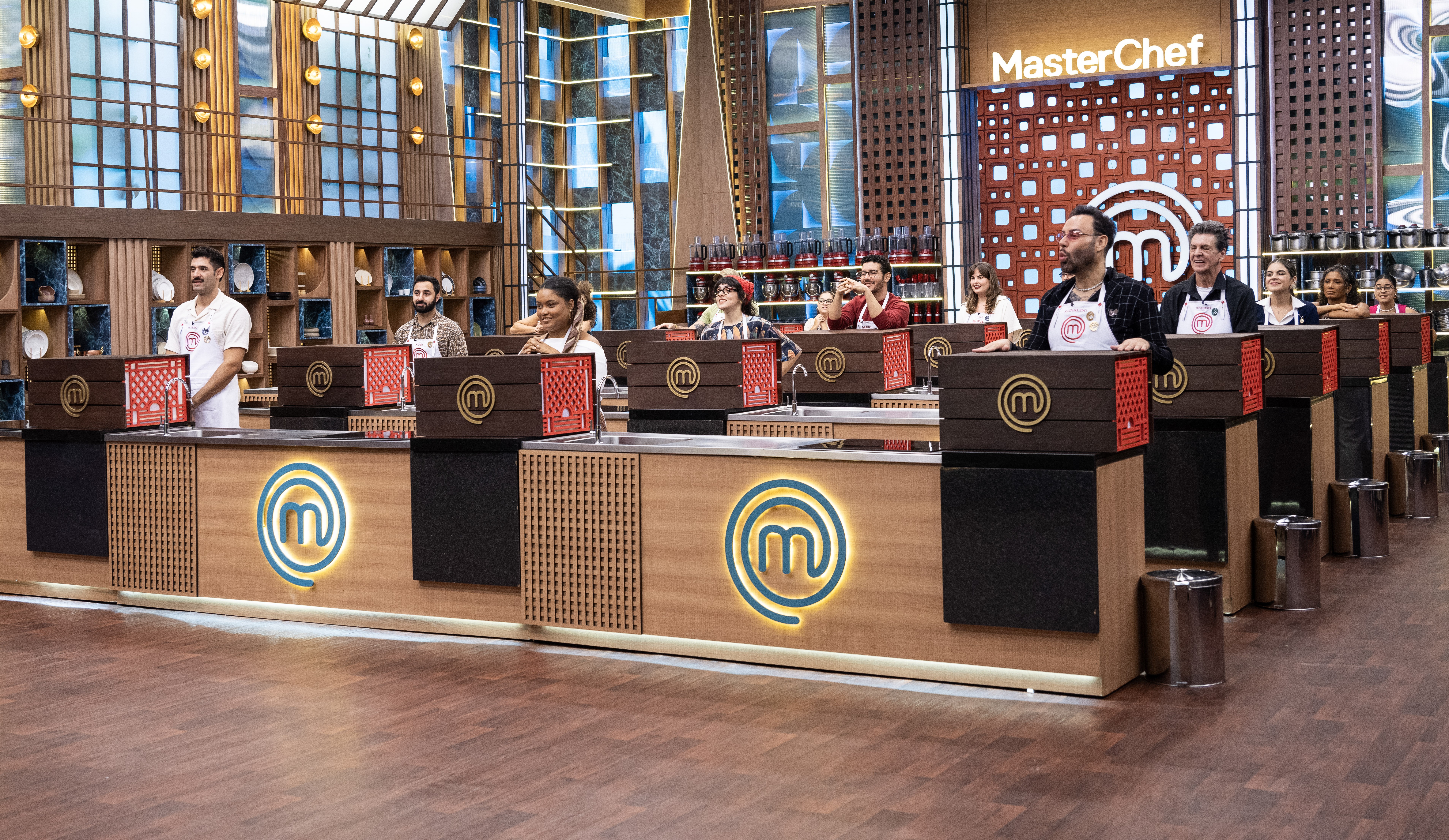 Competidores do MasterChef Brasil, homens e mulheres de diversas etnias, em pé atrás de bancadas de madeira com o logo M em neon azul, no estúdio do programa