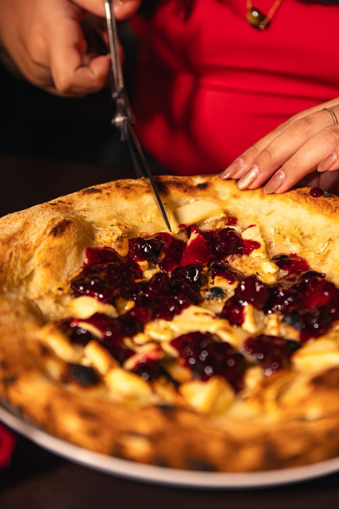 Mãos femininas com unhas claras cortando uma pizza doce com massa crocante, coberta por queijo branco e calda escura de frutas vermelhas