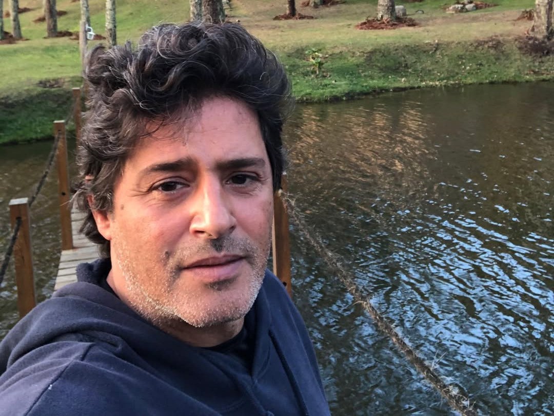 Homem de meia-idade, cabelos escuros e barba rala, usando moletom azul, tira uma selfie em uma ponte de madeira sobre um rio, com árvores ao fundo
