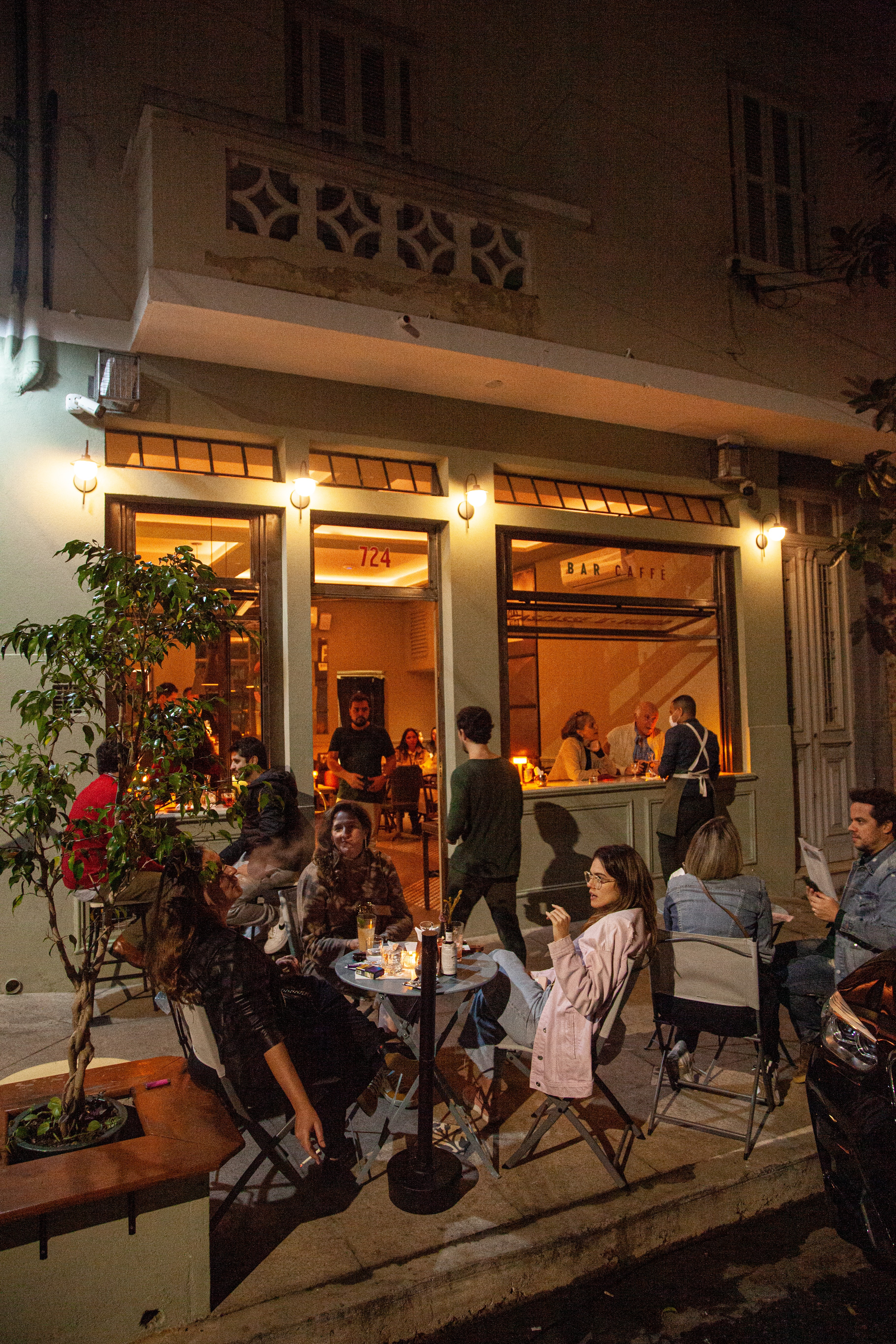 Fachada de bar à noite com pessoas sentadas em mesas na calçada e dentro, iluminado por luzes quentes