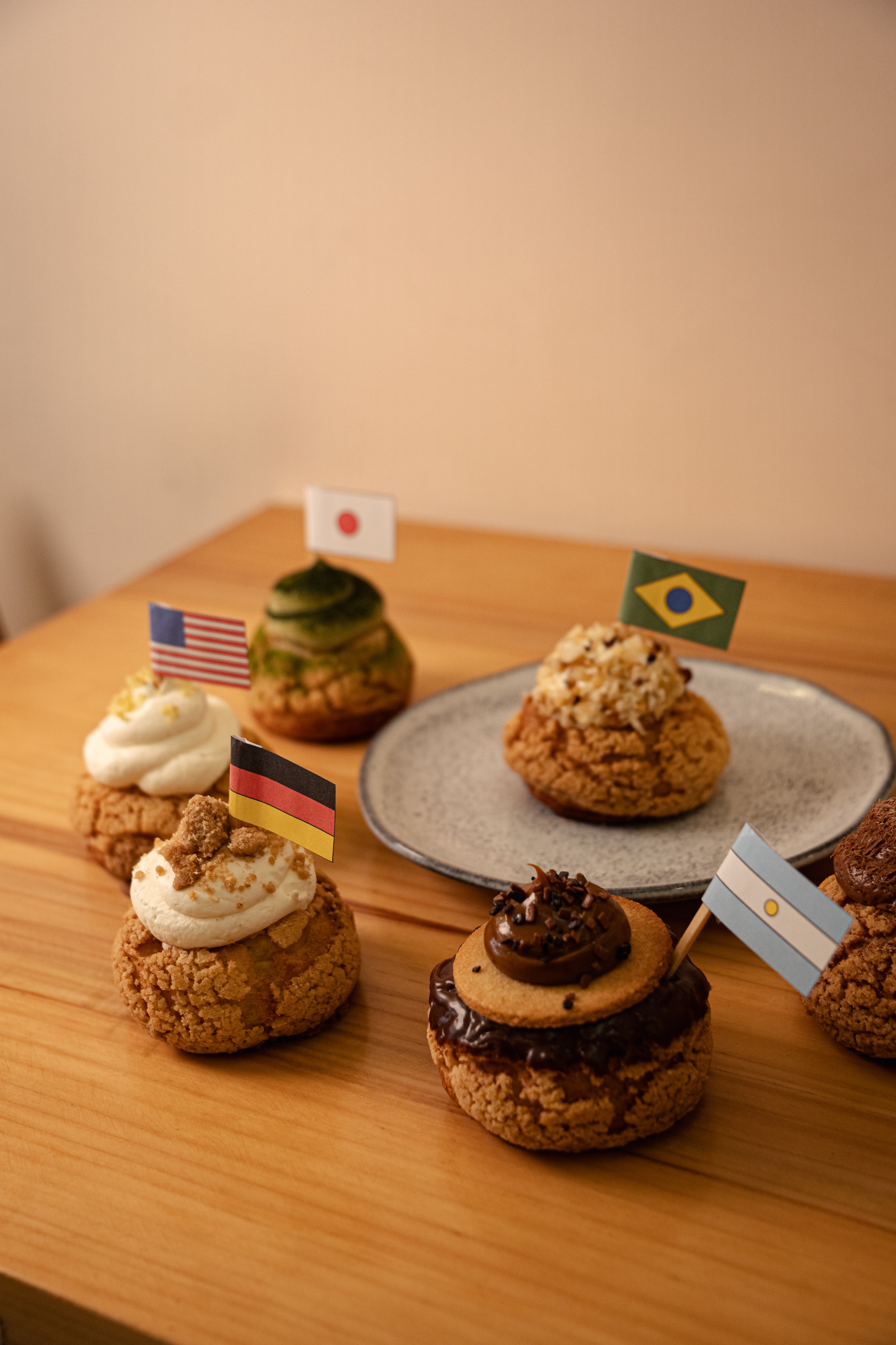 Seis doces redondos com coberturas variadas e pequenas bandeiras de países, como Brasil, Japão e EUA, sobre uma mesa de madeira