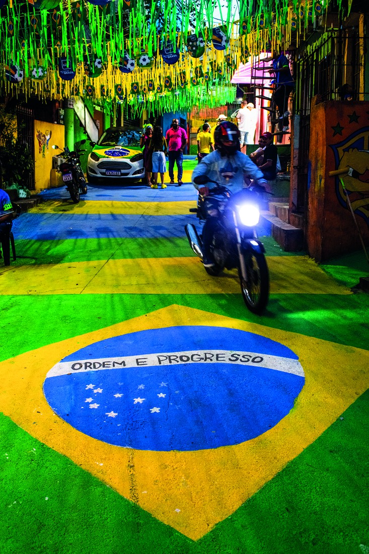 Rua brasileira decorada com as cores da bandeira, verde e amarelo, e faixas azuis. O chão tem um grande círculo azul com a frase ORDEM E PROGRESSO e estrelas brancas. Um motociclista com capacete e jaqueta clara dirige em primeiro plano, com a luz do farol acesa. Pessoas e carros decorados com a bandeira do Brasil aparecem ao fundo, sob enfeites pendurados no teto.