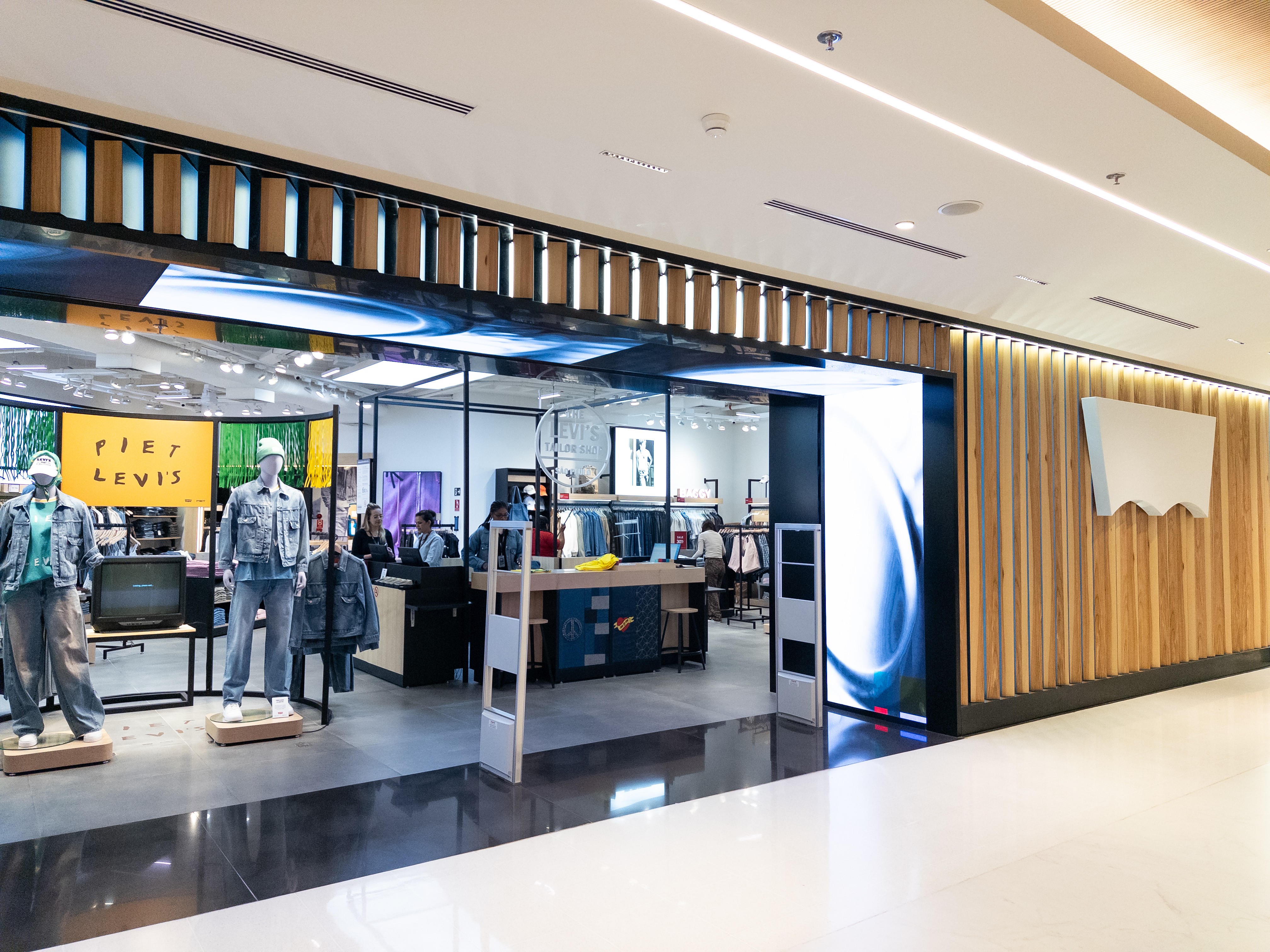Fachada de uma loja Levis em um shopping, com manequins vestindo jeans na vitrine. O interior da loja é visível, com roupas expostas e funcionários. A parede externa tem ripas de madeira e o logotipo da marca em branco.