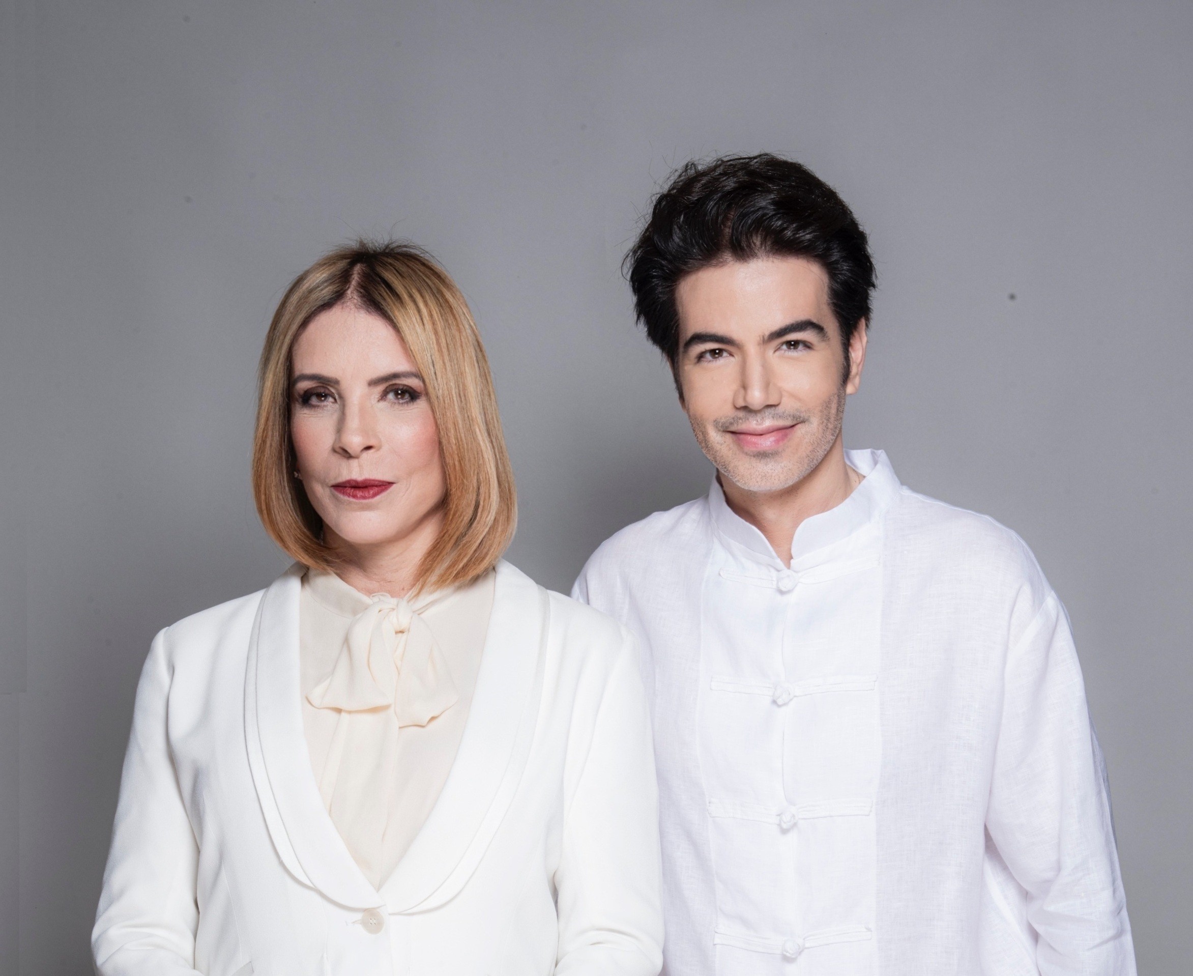 Mulher loira de blazer branco e blusa creme com laço, ao lado de homem de cabelo escuro e camisa branca com botões orientais, ambos sorrindo levemente para a câmera em fundo cinza