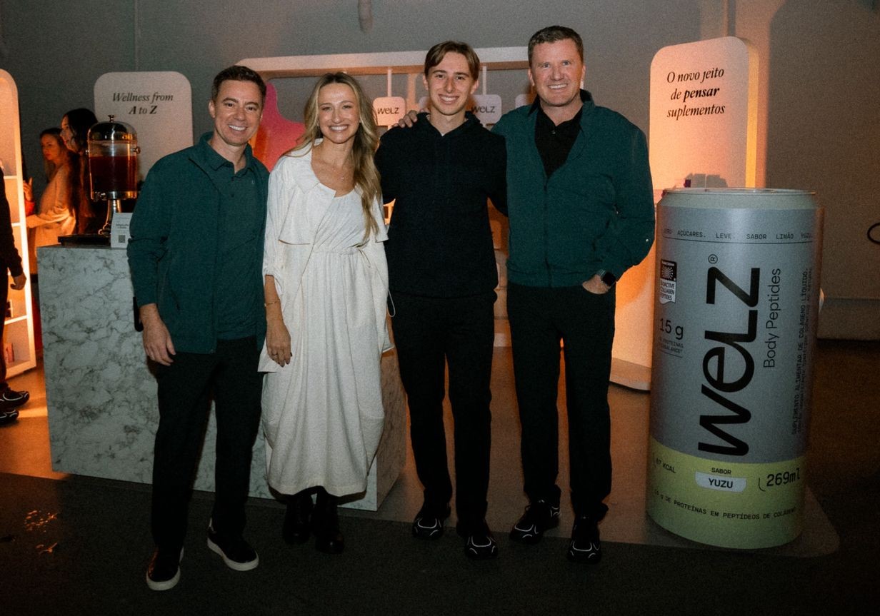 Quatro pessoas sorridentes, três homens e uma mulher, posam em um evento. A mulher veste um vestido branco longo, os homens usam camisas escuras e calças pretas. Ao fundo, um banner branco com Wellness from A to Z e um display com O novo jeito de pensar suplementos. À direita, um grande pote de suplemento Welz Body Peptides