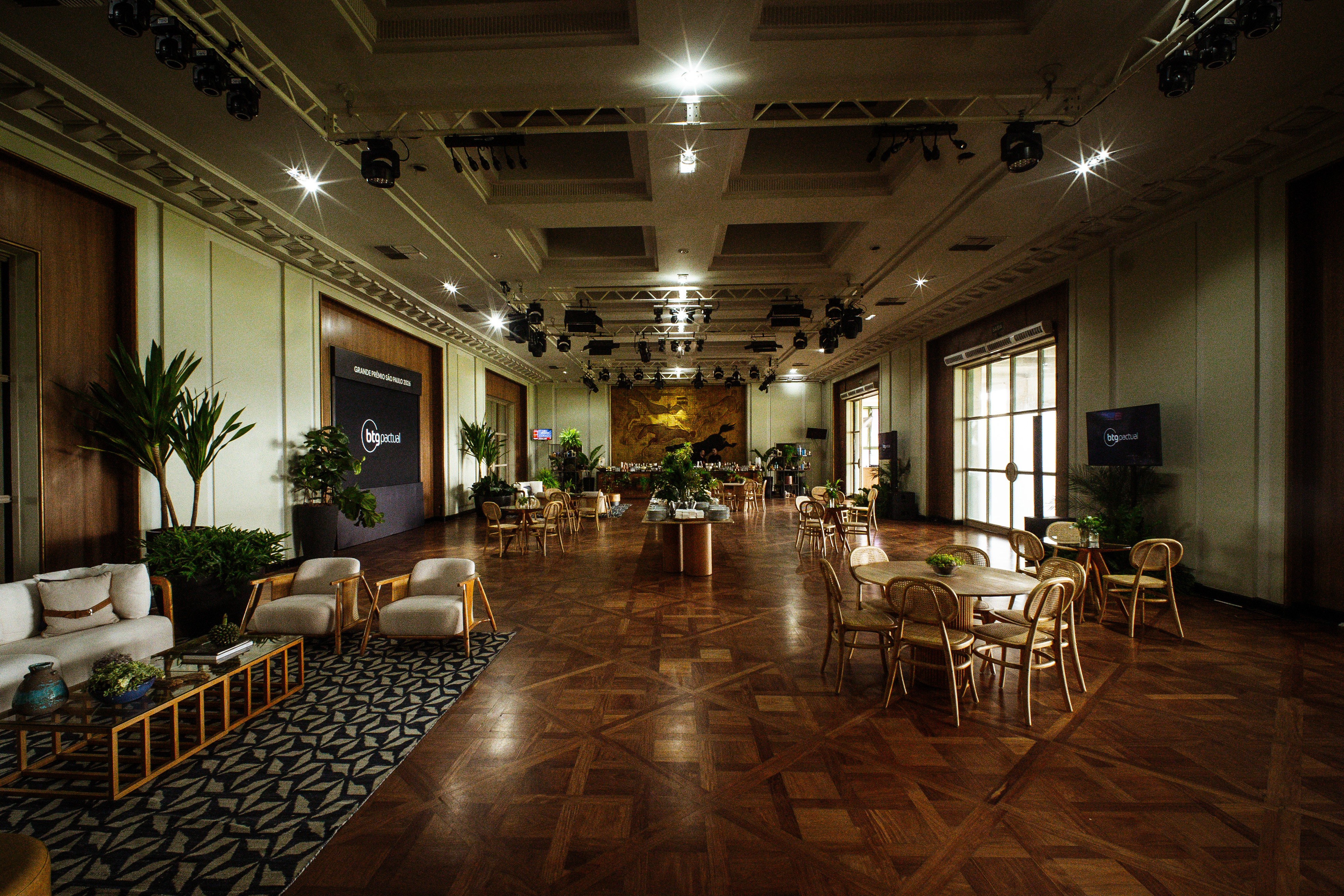 Salão de eventos amplo com piso de madeira, mesas redondas e cadeiras de vime, sofás claros e poltronas, plantas verdes, iluminação focada no teto e telas pretas exibindo o logo bigportal
