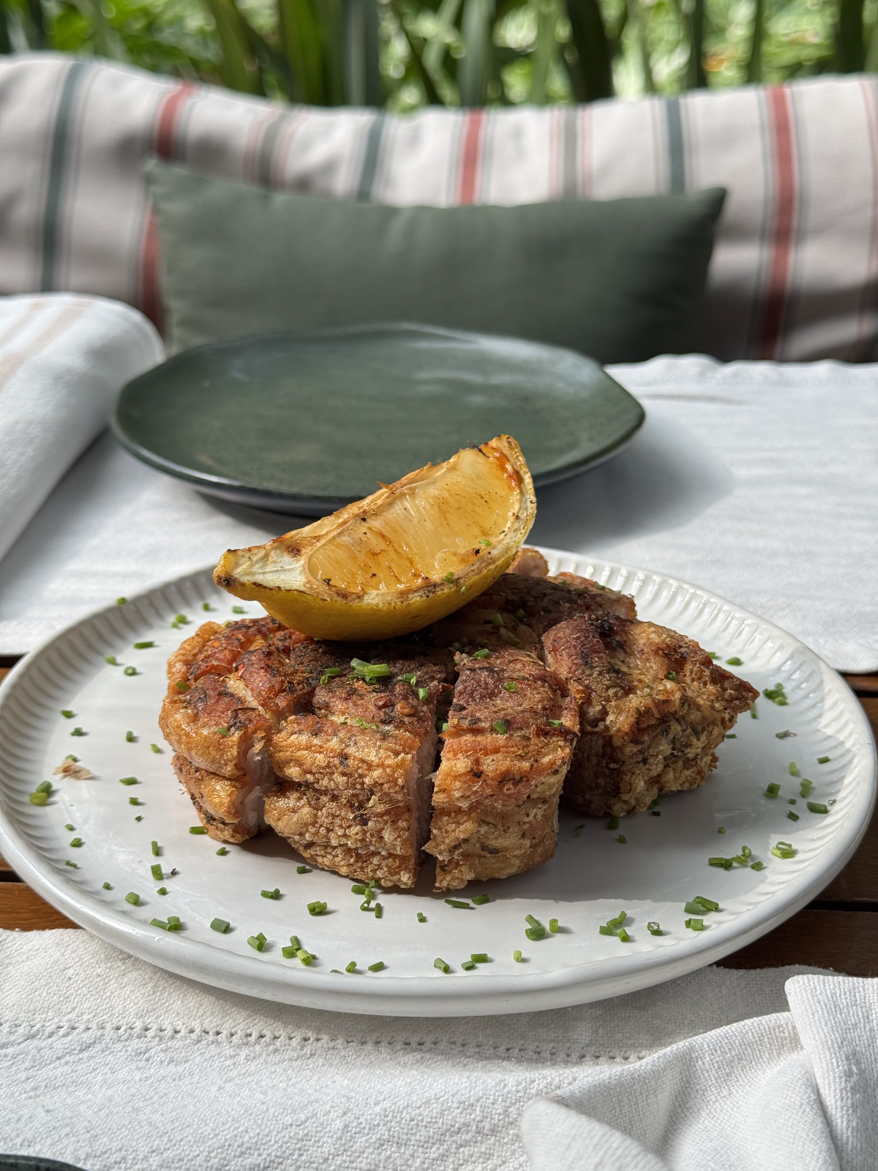 Porção de torresmo crocante em pedaços, polvilhada com cebolinha picada e uma fatia de limão grelhado, servida em prato branco sobre toalha de mesa clara, com um prato vazio e almofadas listradas ao fundo em ambiente externo
