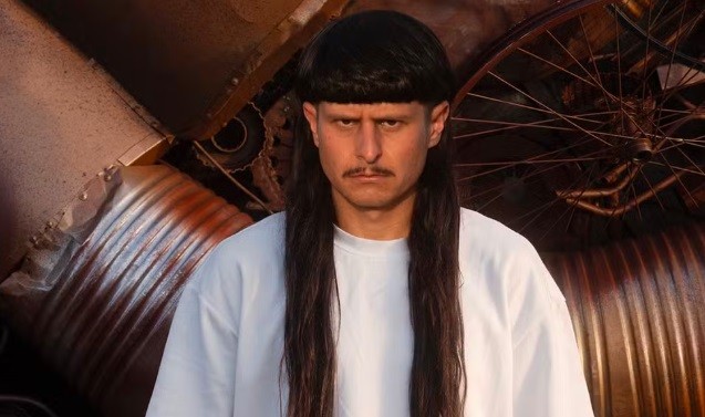 Homem com cabelo comprido e franja reta, bigode fino, expressão séria e testa franzida, vestindo camiseta branca, em frente a um fundo industrial com peças metálicas e uma roda enferrujada