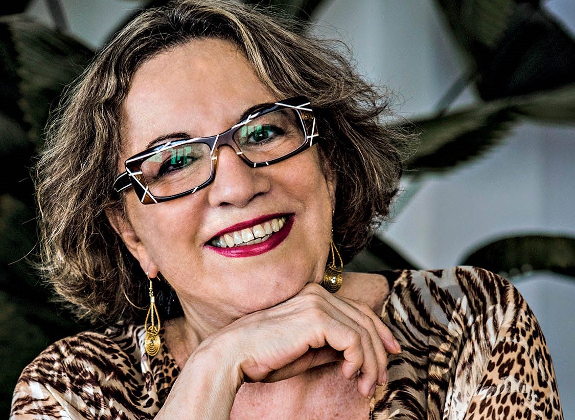 Mulher de meia-idade, cabelos curtos e ondulados, sorri com batom vermelho e óculos modernos de armação preta e branca. Veste blusa estampada de oncinha e brincos dourados, com a mão direita apoiando o queixo. Fundo desfocado com folhagens escuras
