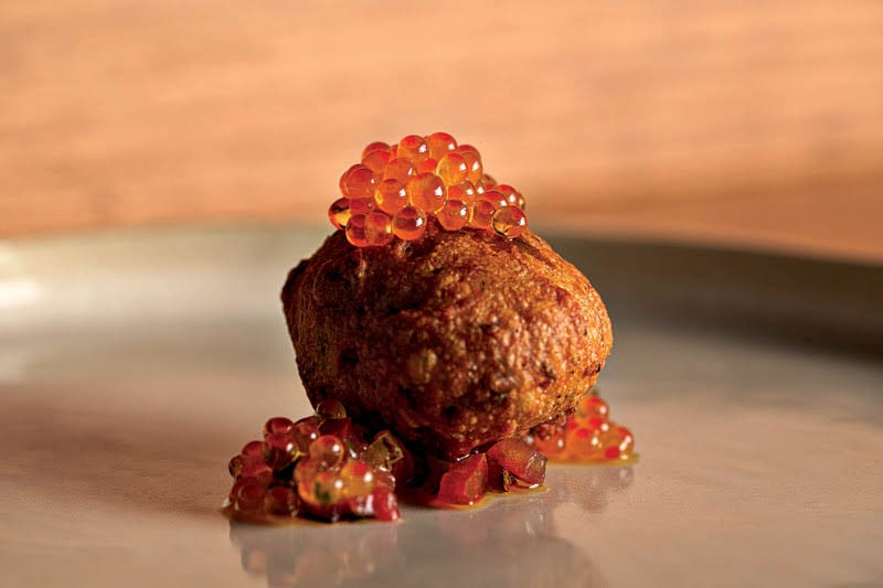 Bolinho frito dourado com cobertura de ovas de salmão alaranjadas e pedaços de tomate picado, servido em prato cinza com fundo marrom claro