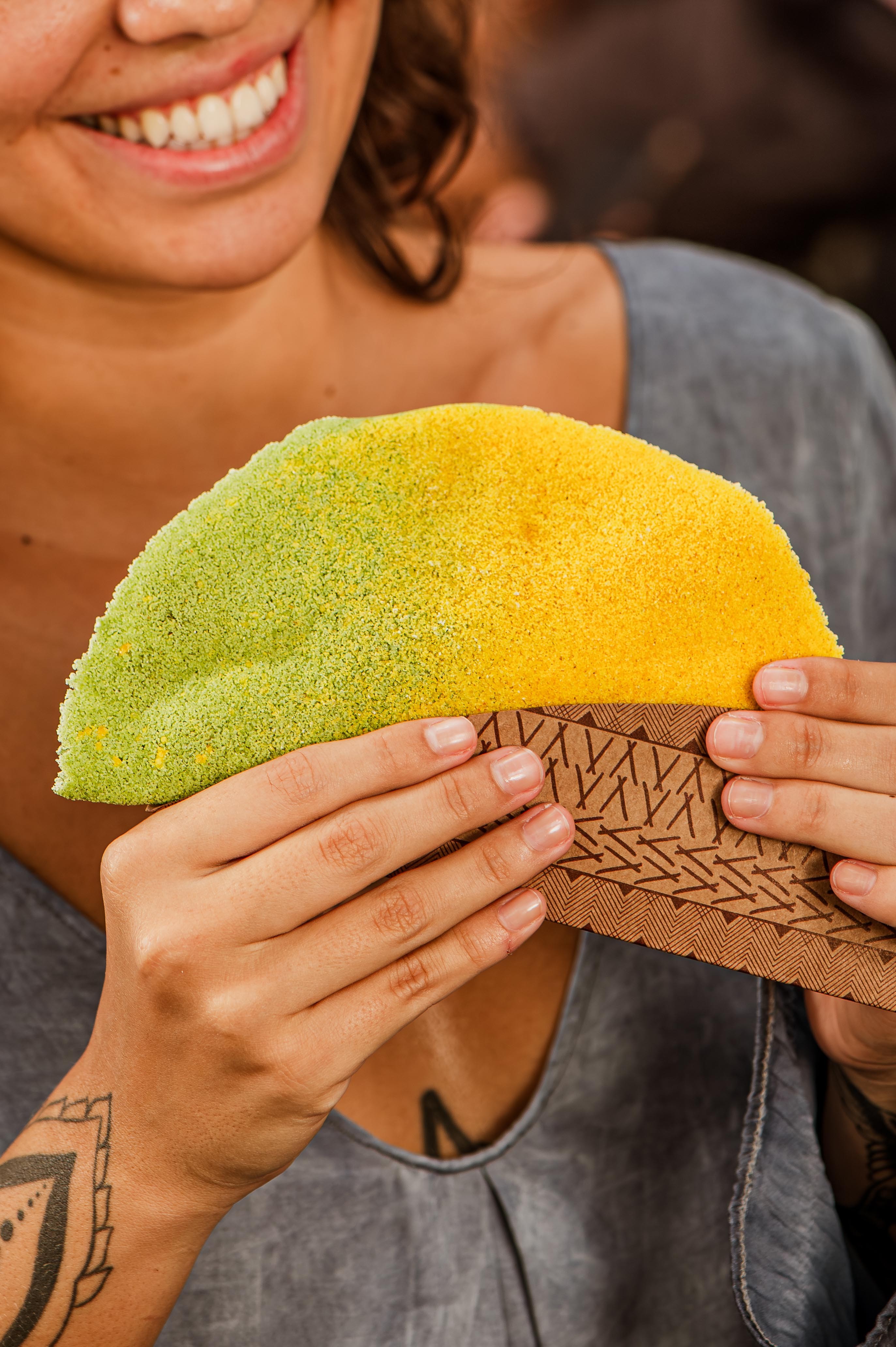 Mulher sorridente segurando uma tapioca colorida, metade verde e metade amarela, com as mãos em um suporte de papel marrom estampado