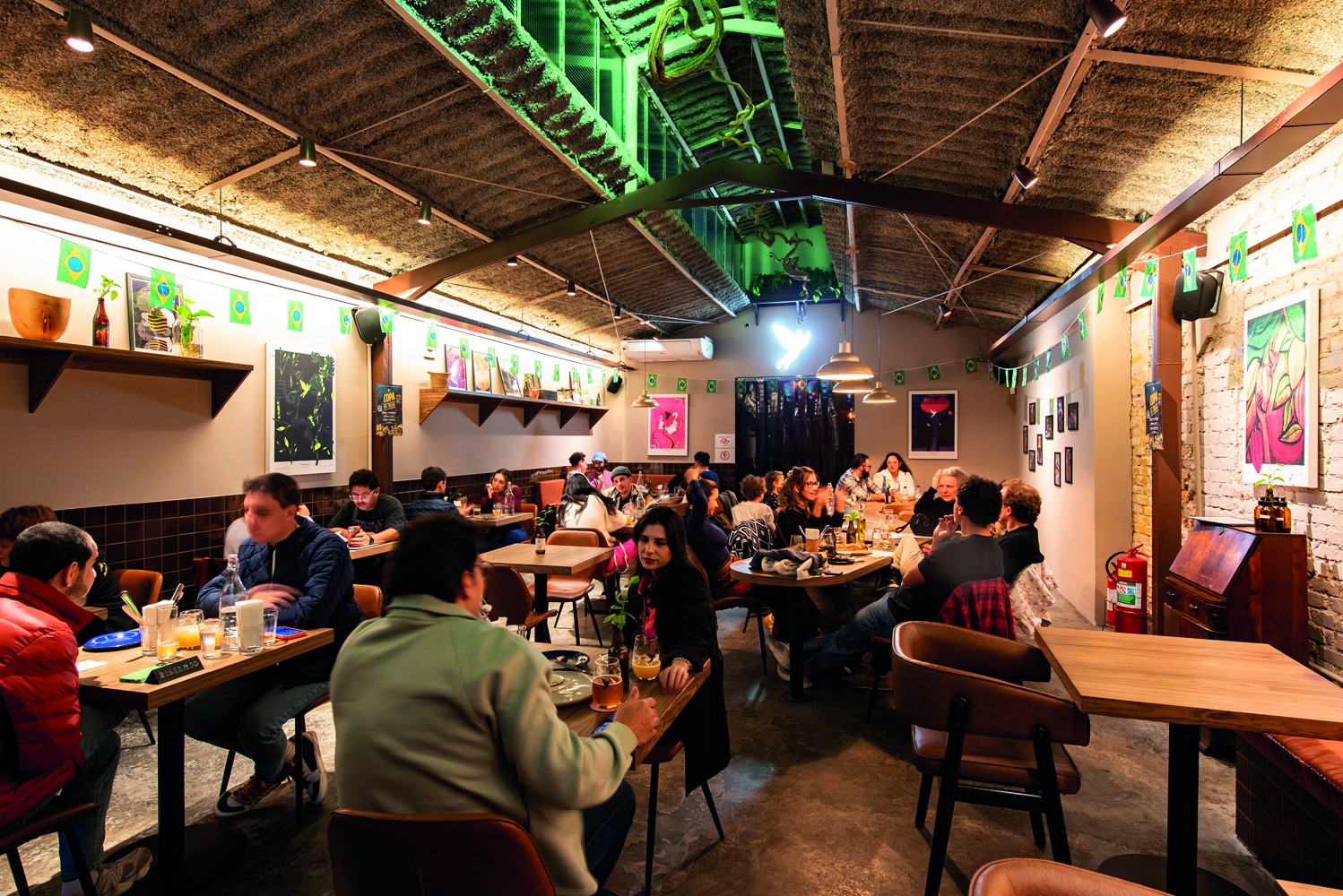 Pessoas sentadas em mesas de madeira em um bar ou restaurante com teto de palha e iluminação quente. As paredes são decoradas com quadros e bandeirinhas verdes.
