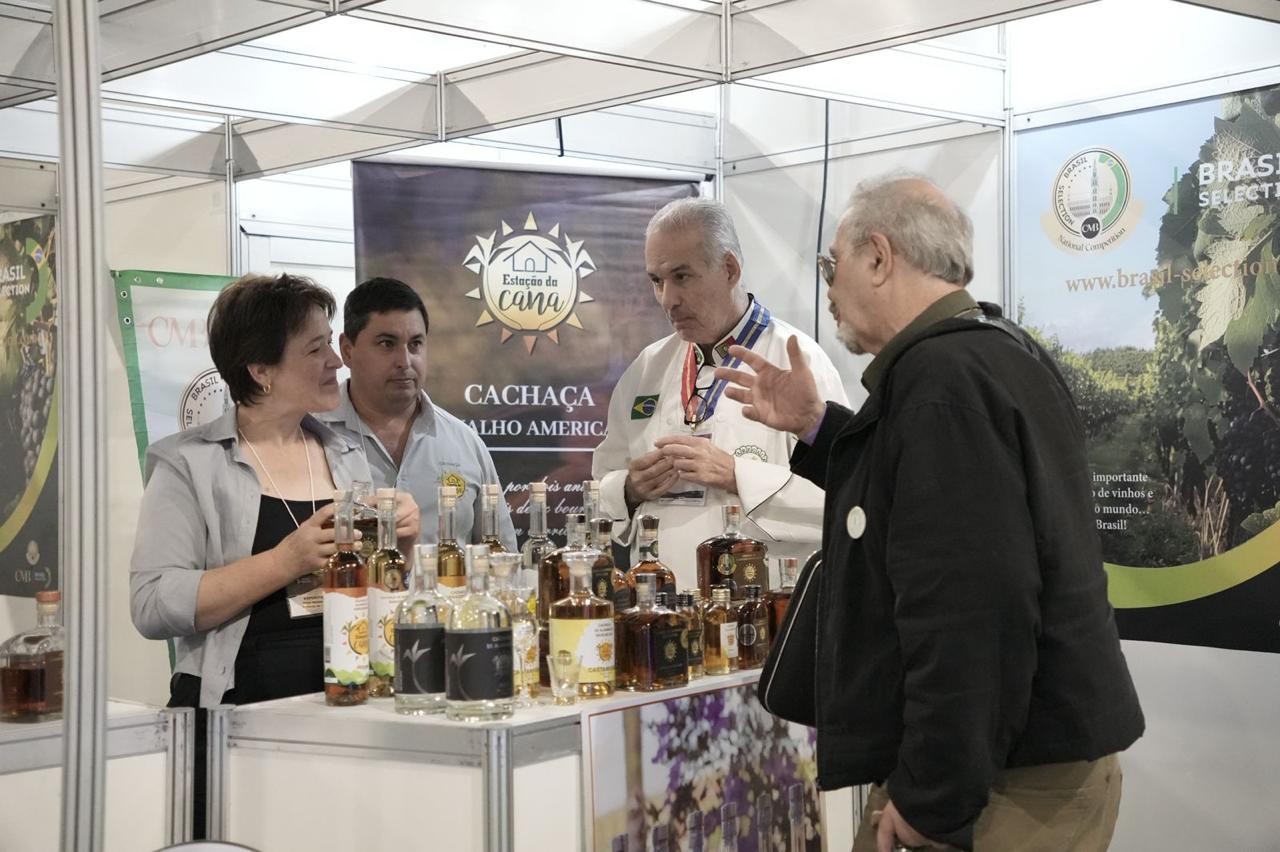 Quatro pessoas em um estande de exposição. Uma mulher de blusa cinza e um homem de camisa azul clara observam garrafas de cachaça. Um homem de jaleco branco e medalhas no pescoço conversa com um homem de casaco preto, que gesticula com a mão direita. Ao fundo, banners de Estação da Cana e Brasil Selection