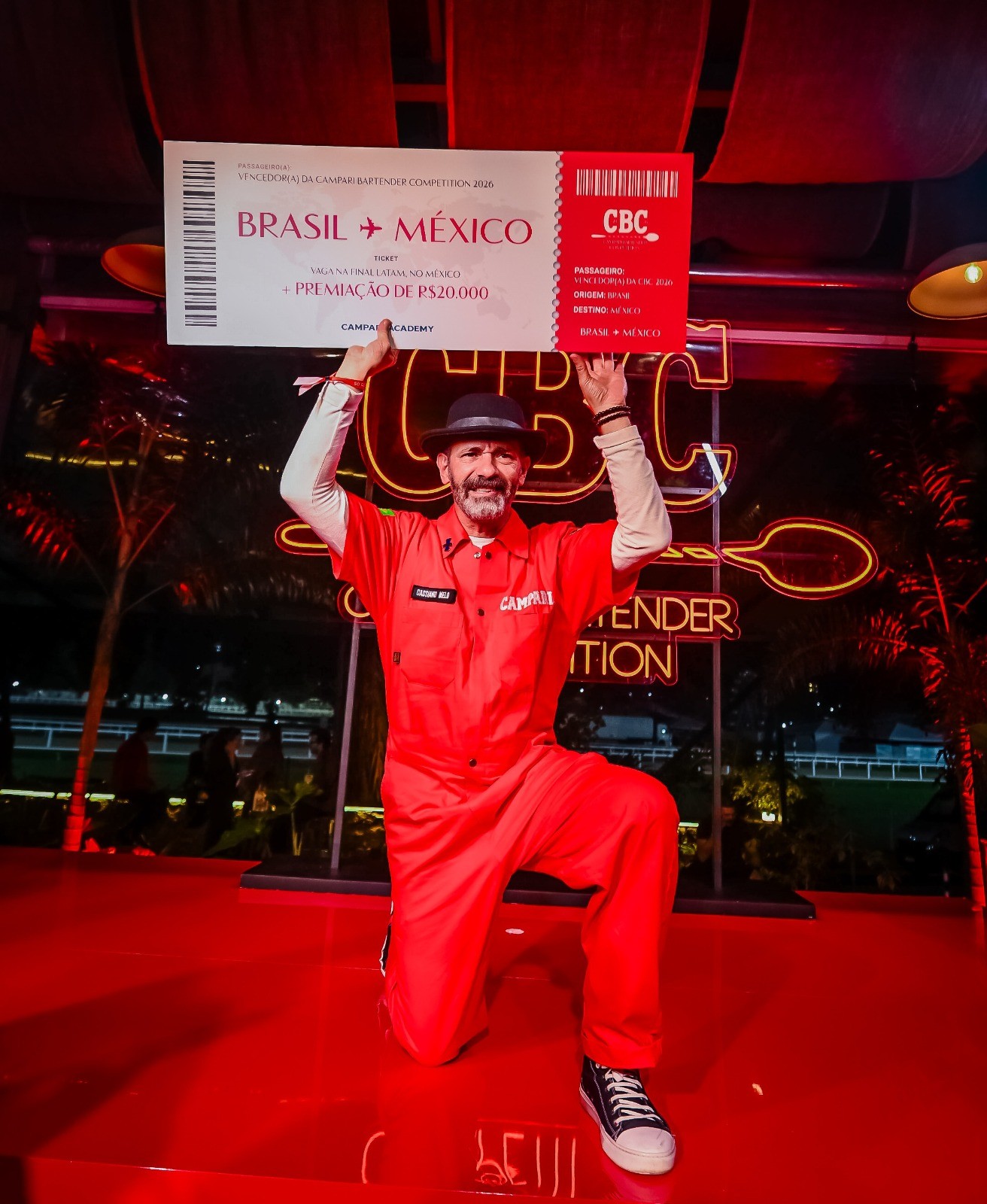 Homem barbudo de chap&eacute;u e macac&atilde;o vermelho, ajoelhado, ergue um bilhete gigante de avi&atilde;o com destino Brasil-M&eacute;xico e premia&ccedil;&atilde;o de R.000, em palco vermelho iluminado por luzes neon
