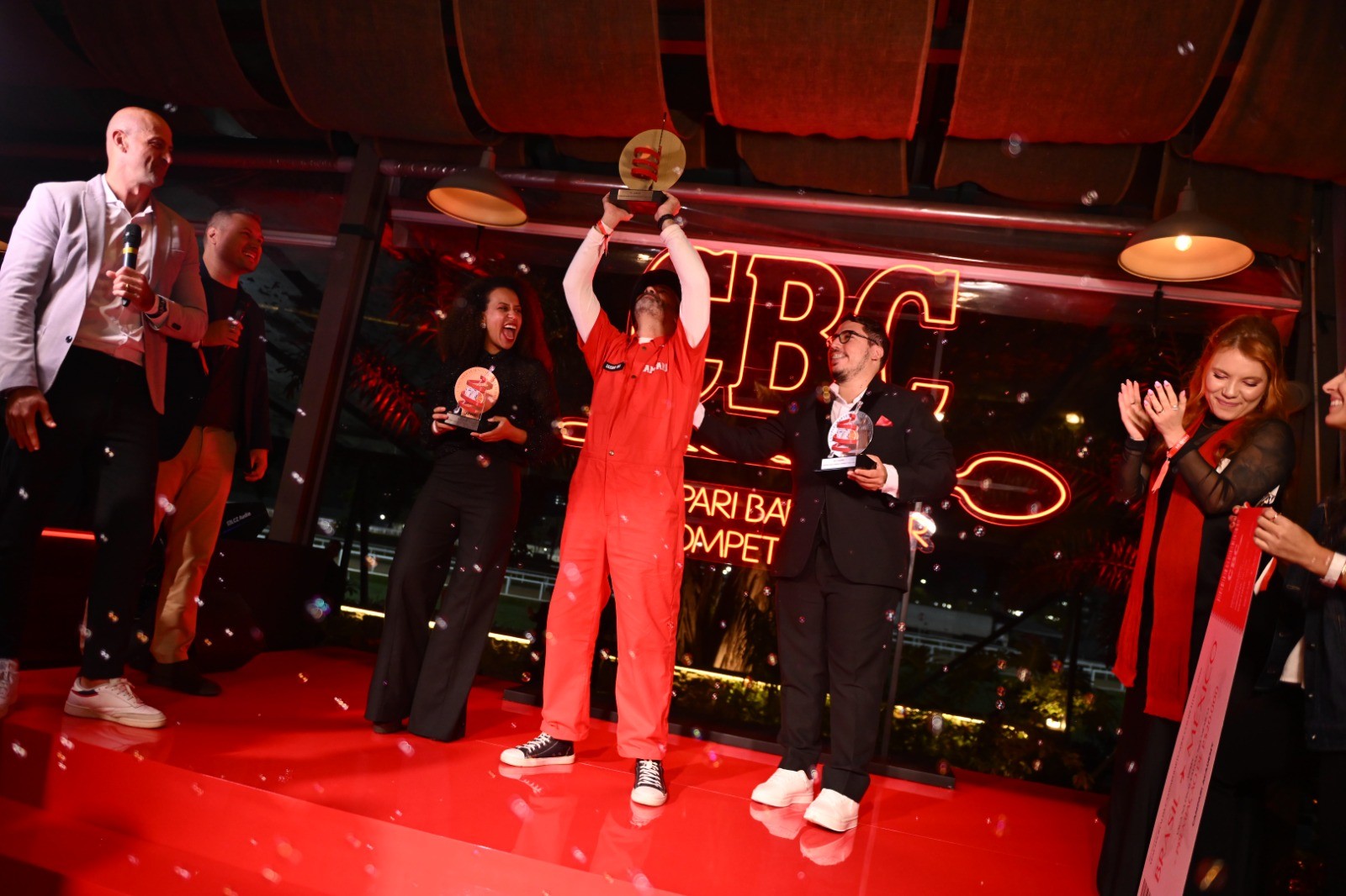Pessoas em um palco vermelho com confetes caindo. Um homem de macac&atilde;o vermelho levanta um trof&eacute;u dourado, enquanto outros seguram pr&ecirc;mios e aplaudem.