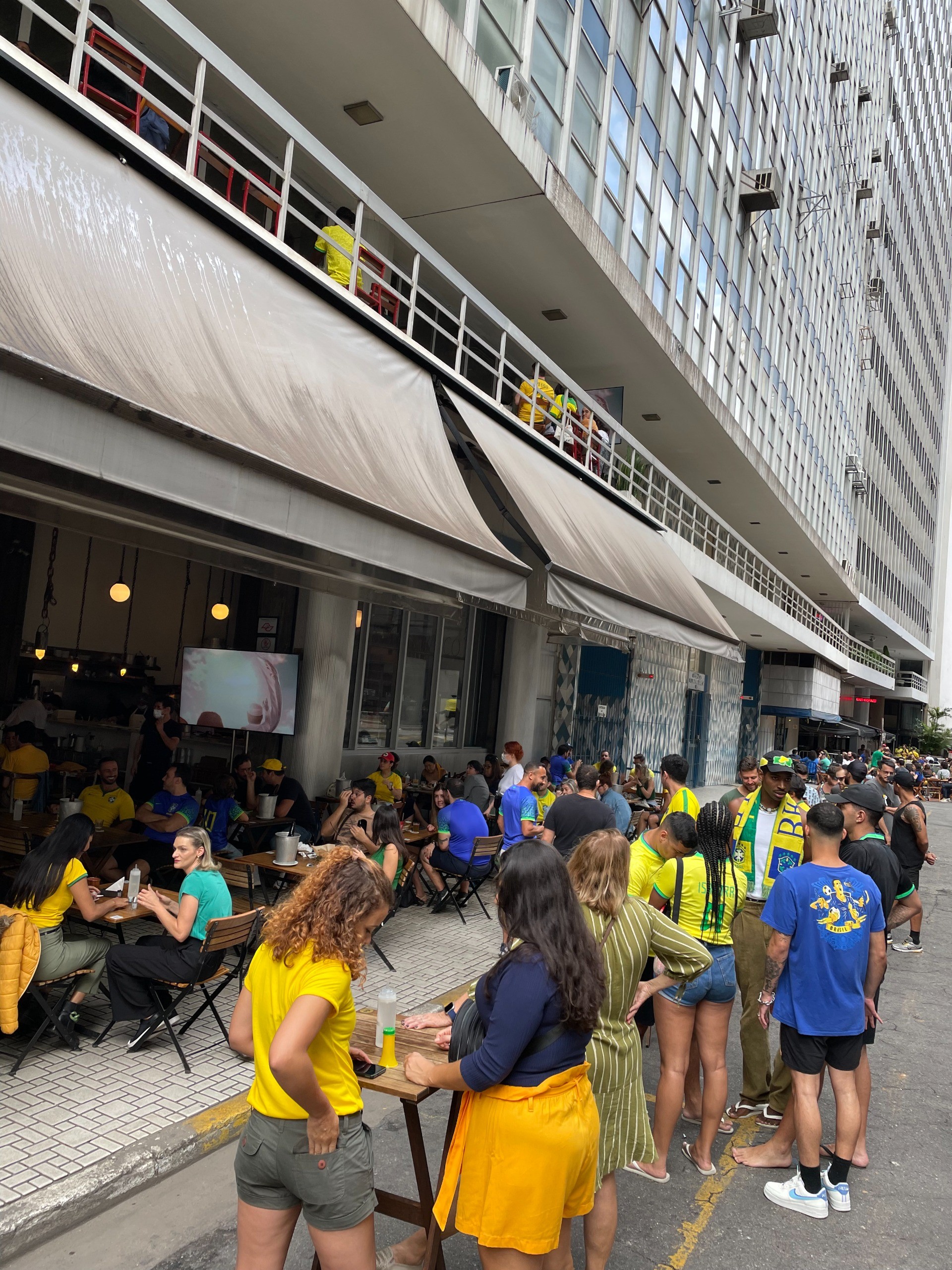 Pessoas vestindo roupas nas cores da bandeira brasileira, como amarelo e azul, assistem a um jogo de futebol em um bar com mesas na calçada e em uma varanda, sob um prédio alto com muitas janelas