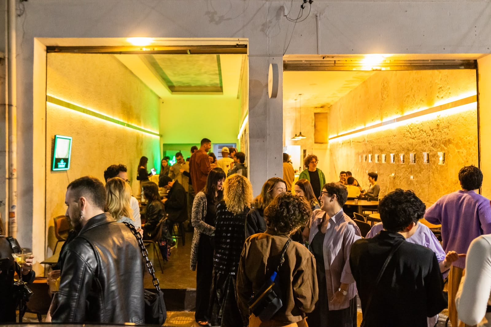 Pessoas conversando e socializando em um bar ou evento noturno, com iluminação amarela e verde neon nas paredes, criando uma atmosfera vibrante