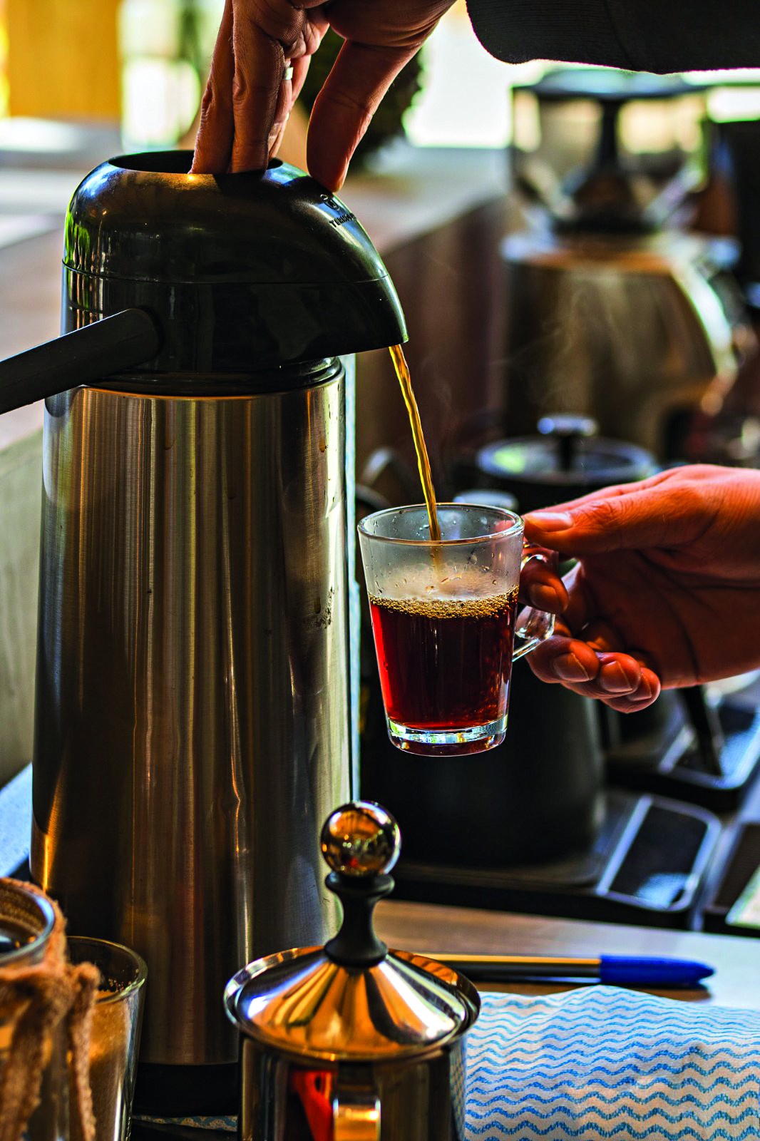 Uma pessoa serve café quente de uma garrafa térmica de inox em uma xícara de vidro transparente, segurada pela outra mão. O café forma um fio dourado, e a xícara já contém um pouco da bebida escura, com vapor subindo. Ao fundo, outros utensílios de cozinha desfocados.