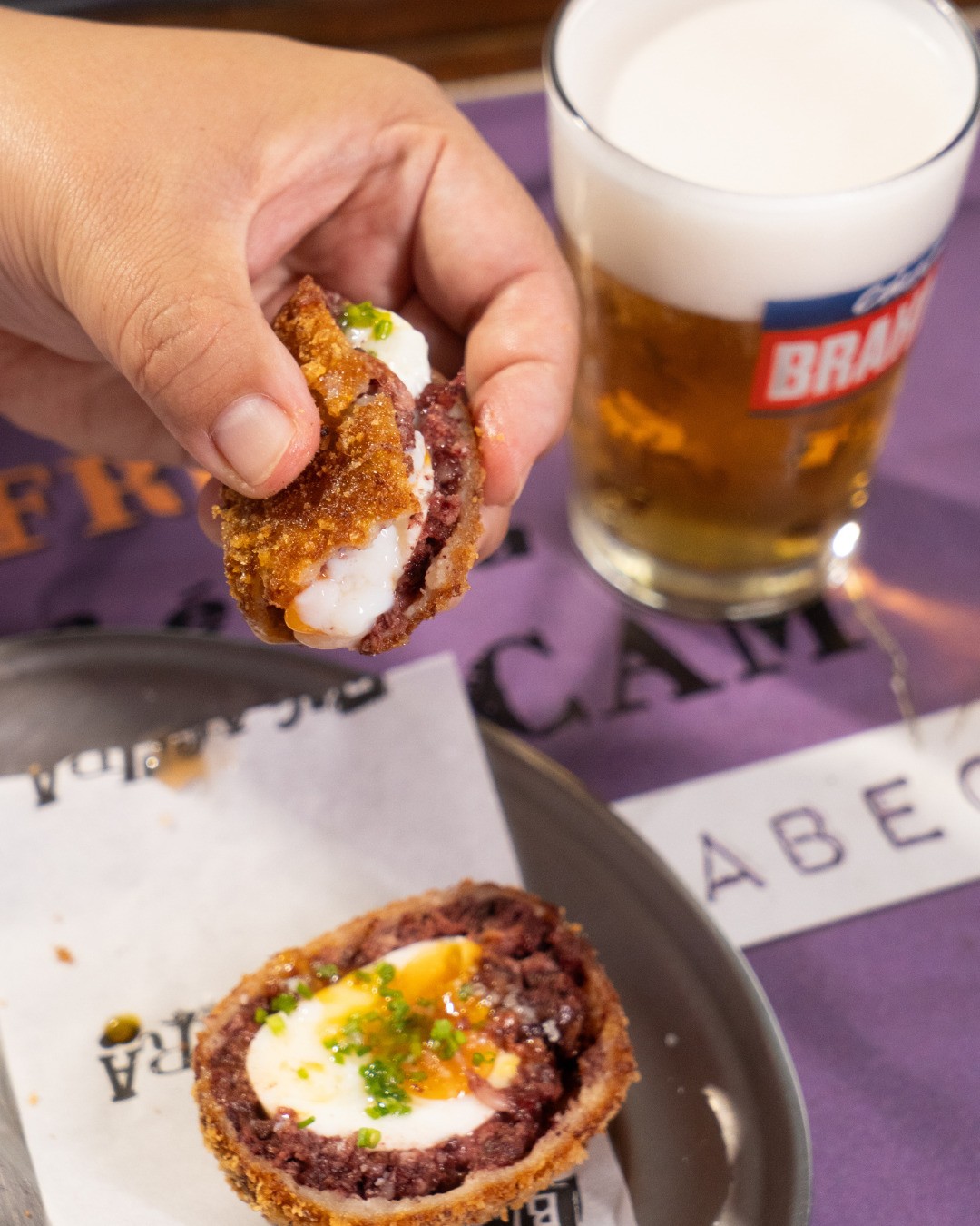 Mão segura um bolinho de carne frito com recheio branco, enquanto outro bolinho com ovo cozido e cheiro-verde repousa sobre papel. Ao fundo, um copo de cerveja e um tecido roxo com letras.