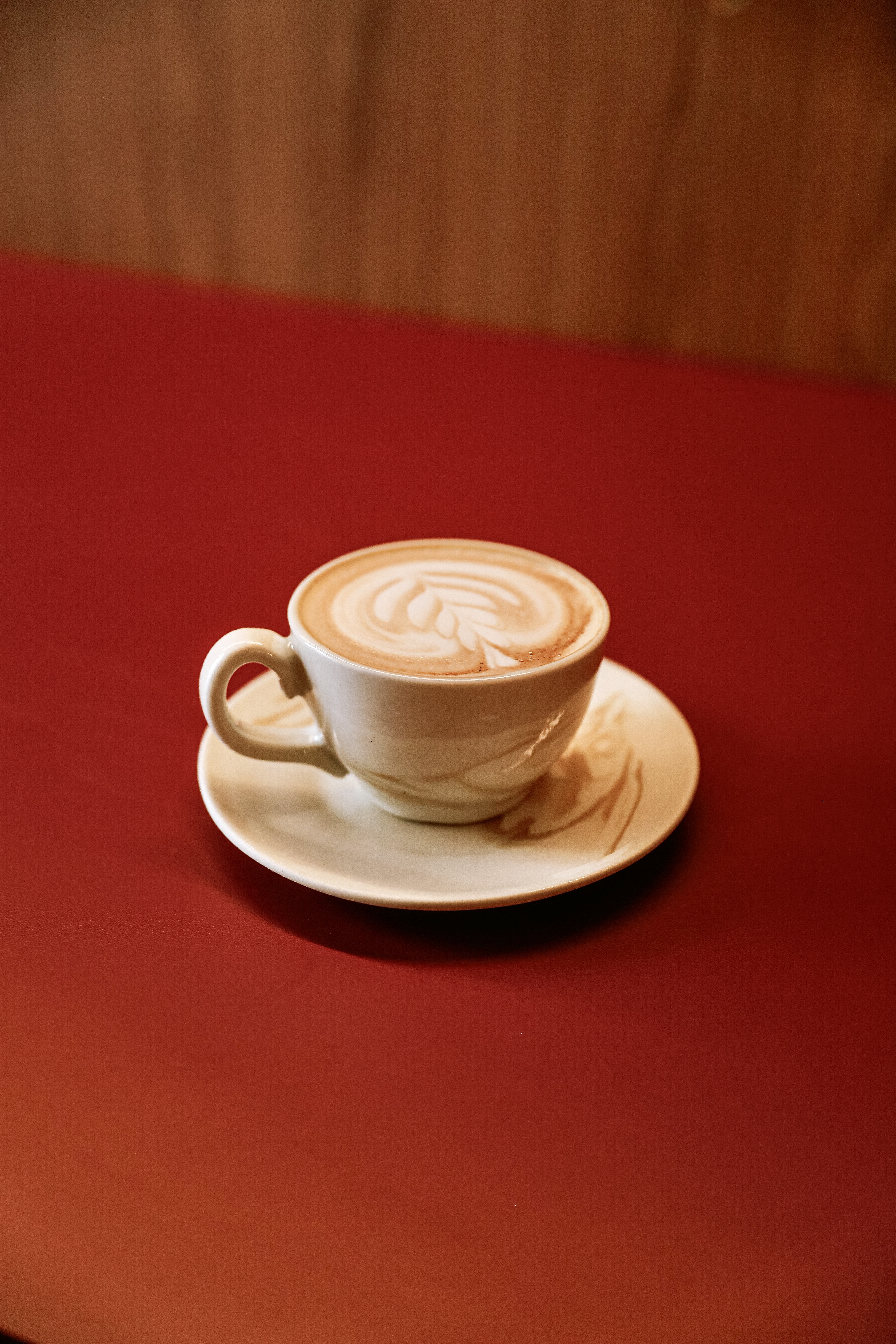 Xícara branca de café latte com arte de folha na espuma, sobre um pires branco, em uma mesa vermelha