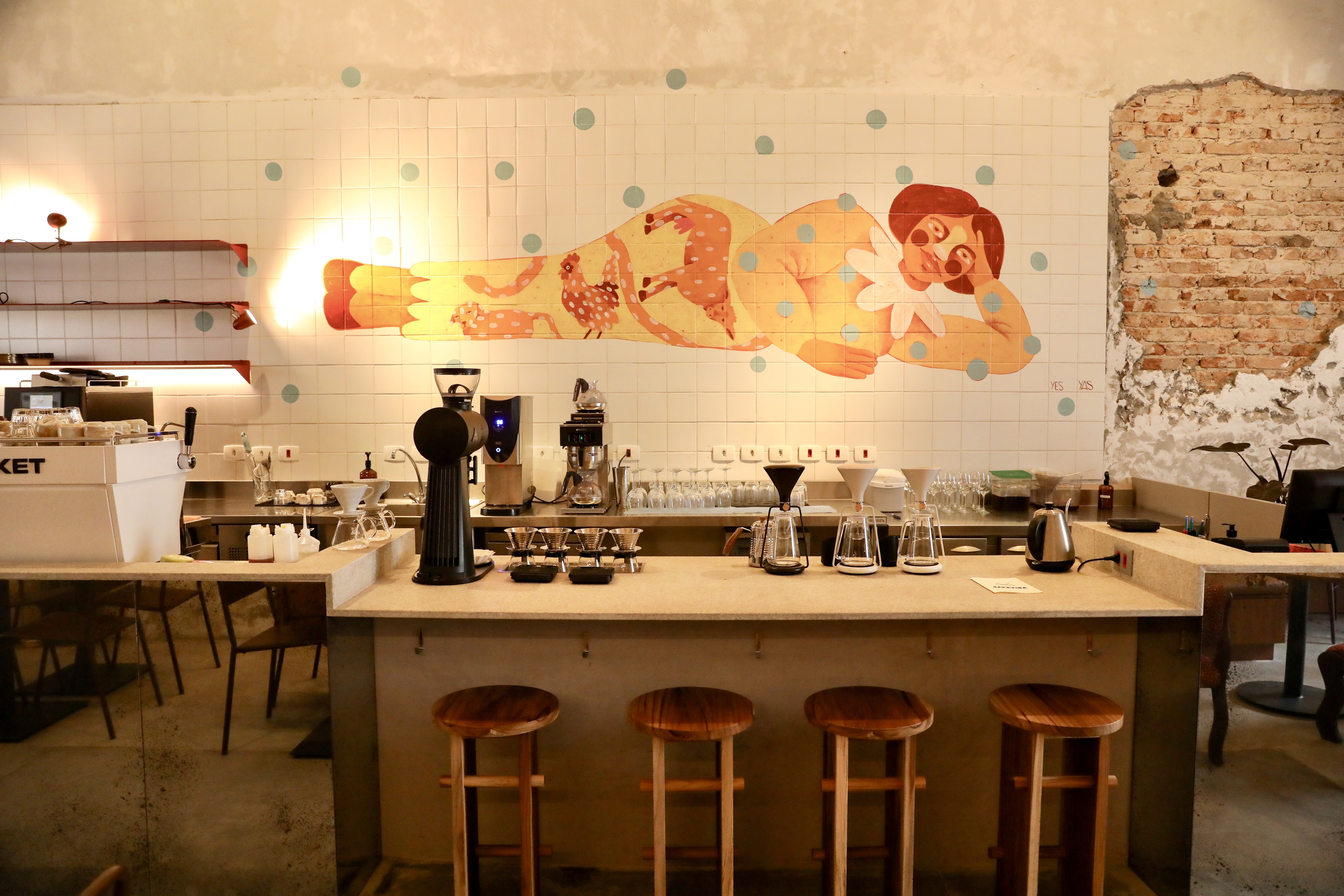Interior de uma cafeteria com balcão de madeira clara e quatro bancos altos de madeira. Ao fundo, uma parede de azulejos brancos com um mural de uma mulher deitada, em tons de amarelo e vermelho, e pontos azuis. Equipamentos de café e copos estão sobre o balcão. À direita, uma parede de tijolos expostos e descascados.