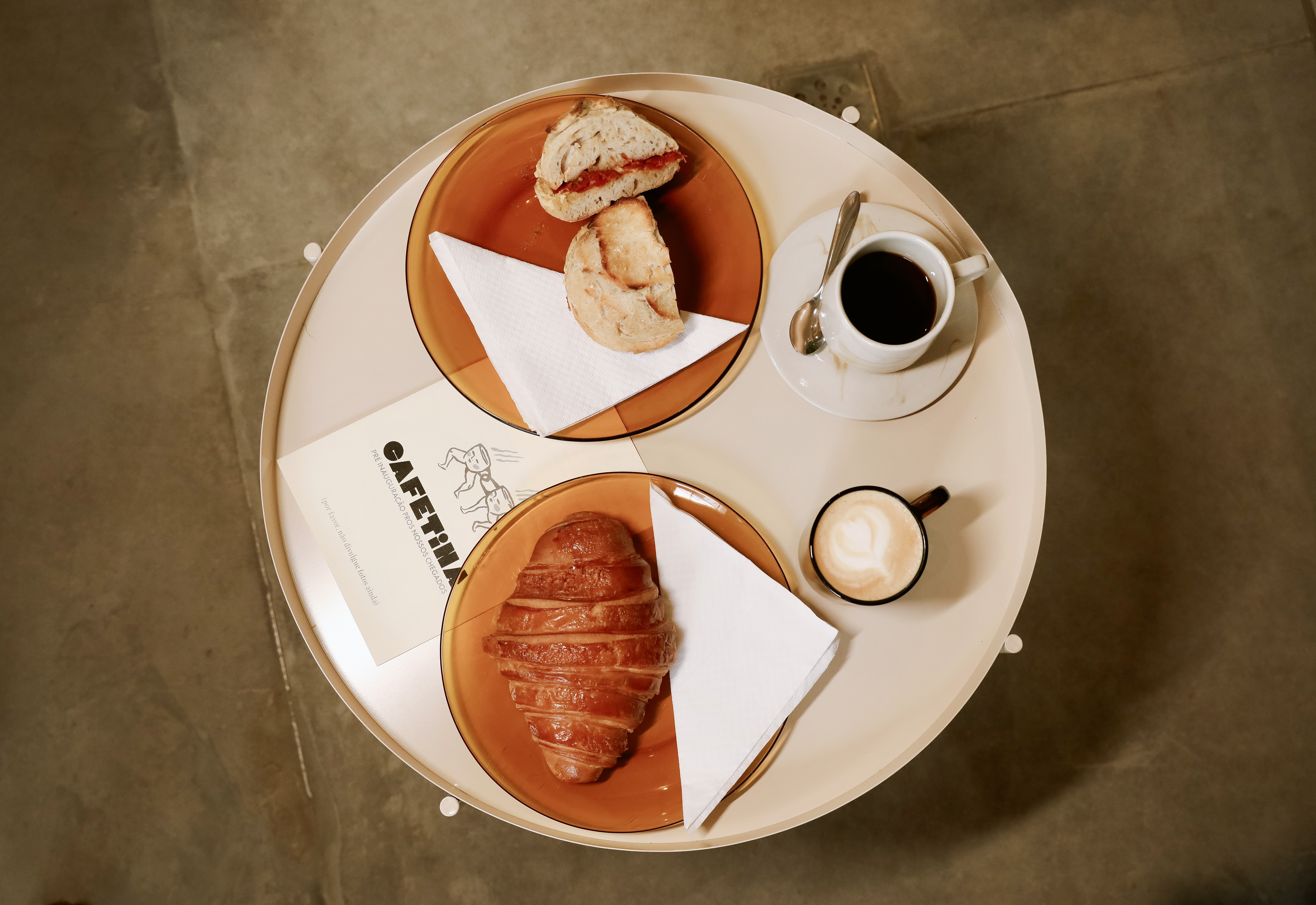 Vista aérea de uma mesa redonda clara com café e lanches. Há um pão com recheio vermelho e outro pão simples em um prato, um croissant fatiado em outro, uma xícara de café preto e uma de cappuccino com arte de coração, e um cardápio CAFETINA