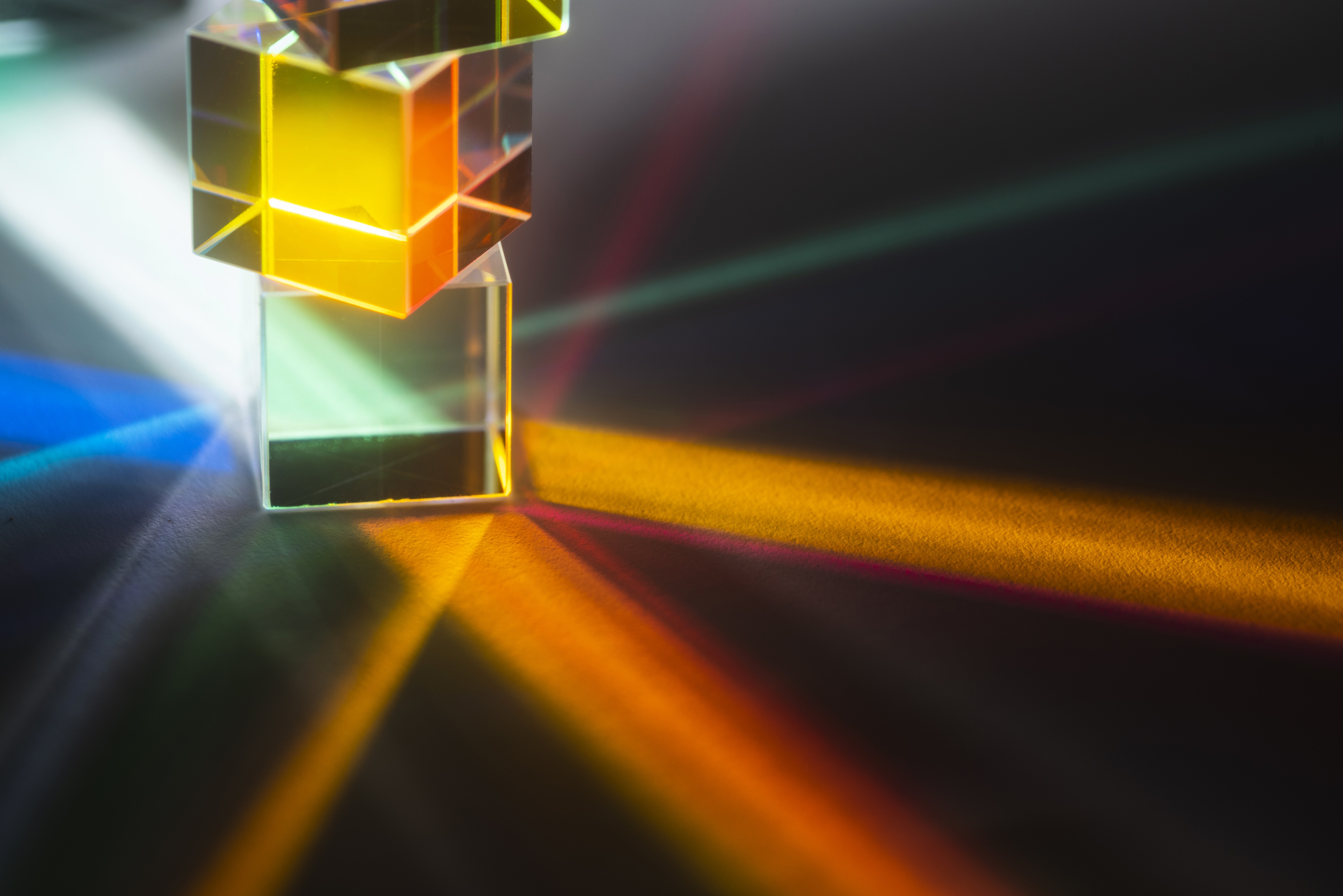 Dois cubos de vidro transparentes empilhados, o superior com reflexos amarelos e laranjas, o inferior com reflexos verdes. Raios de luz coloridos, como azul, amarelo, laranja e vermelho, se espalham pelo fundo escuro, criando um efeito de refração