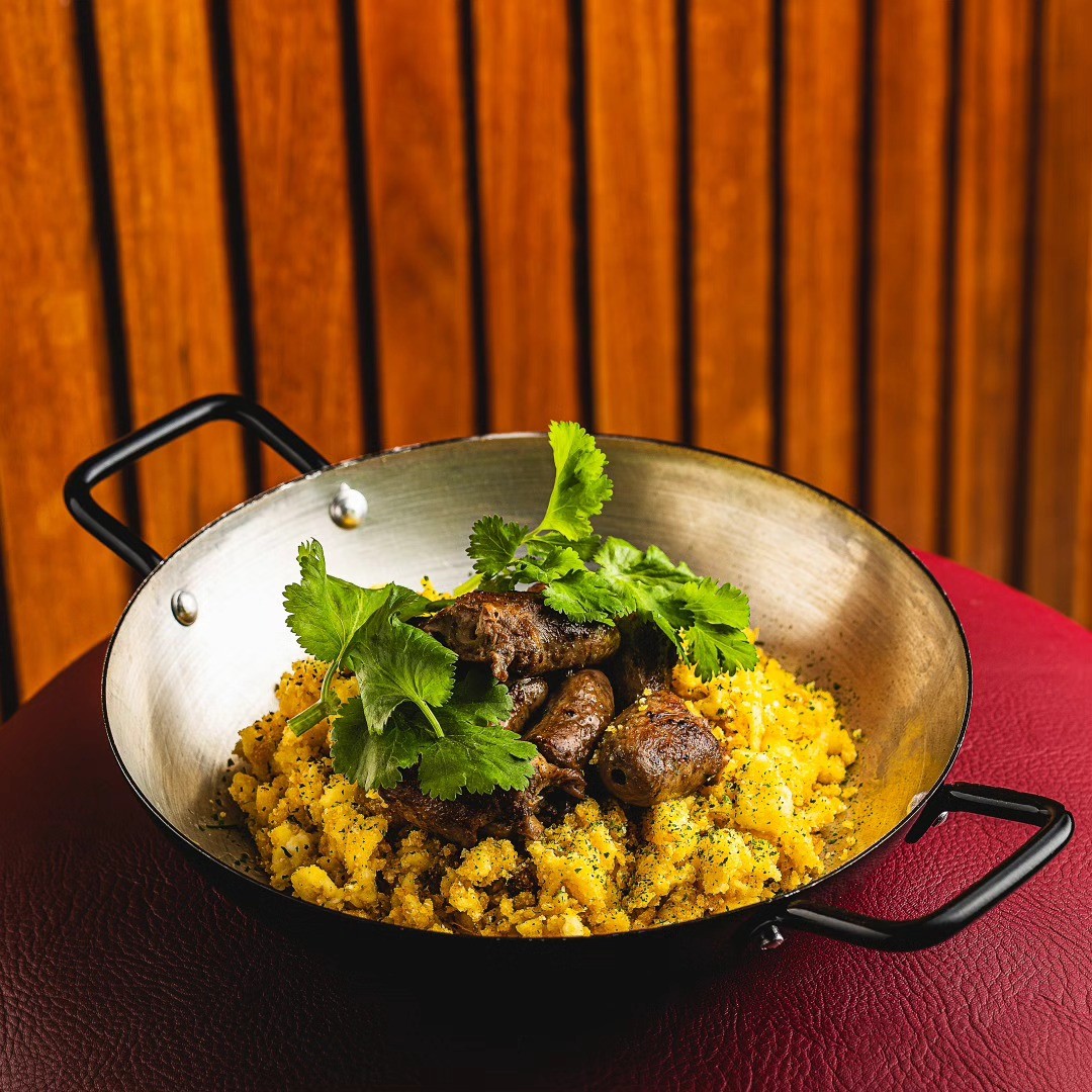 Porção de carne de sol com farofa amarela e folhas de coentro fresco, servida em frigideira de metal com alças pretas sobre superfície vermelha, com fundo de madeira vertical