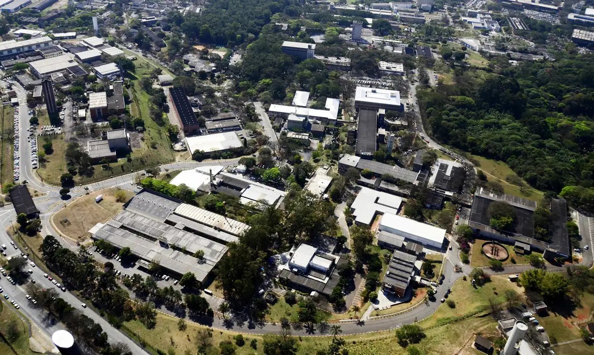 Vista aérea de um campus universitário com diversos edifícios de telhados cinzas e brancos, cercados por áreas verdes e árvores. Ruas asfaltadas com carros estacionados e em movimento cortam o campus, conectando os prédios. Ao fundo, uma área densa de mata.