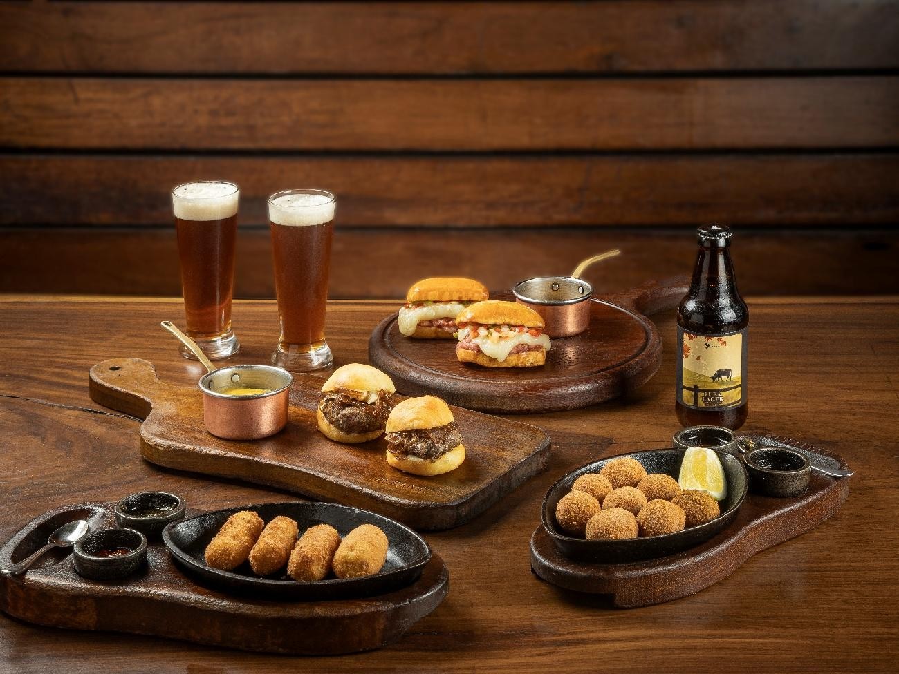 Mesa de madeira com dois copos de cerveja, uma garrafa de cerveja, mini hambúrgueres, croquetes e molhos em tábuas de servir