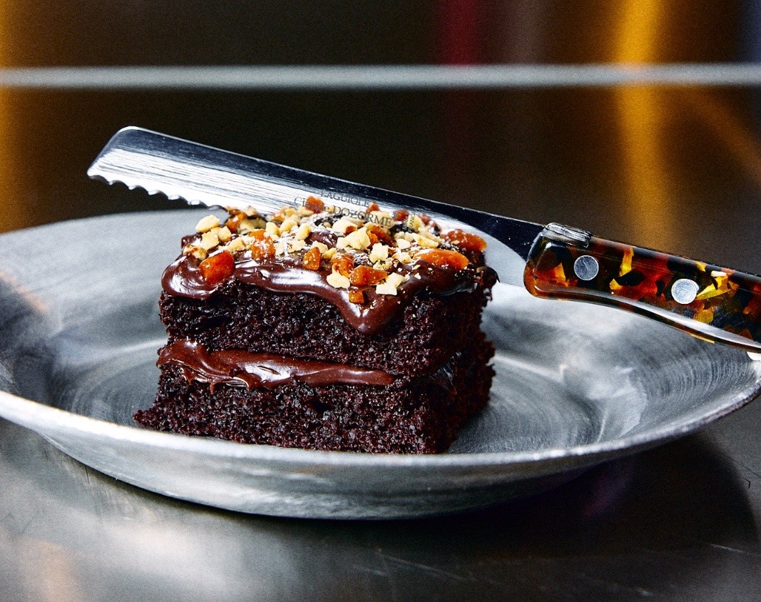 Fatia de bolo de chocolate com duas camadas, cobertura de ganache escorrendo e granulado de nozes e frutas secas, servida em um prato de metal com uma faca de cabo marmorizado apoiada
