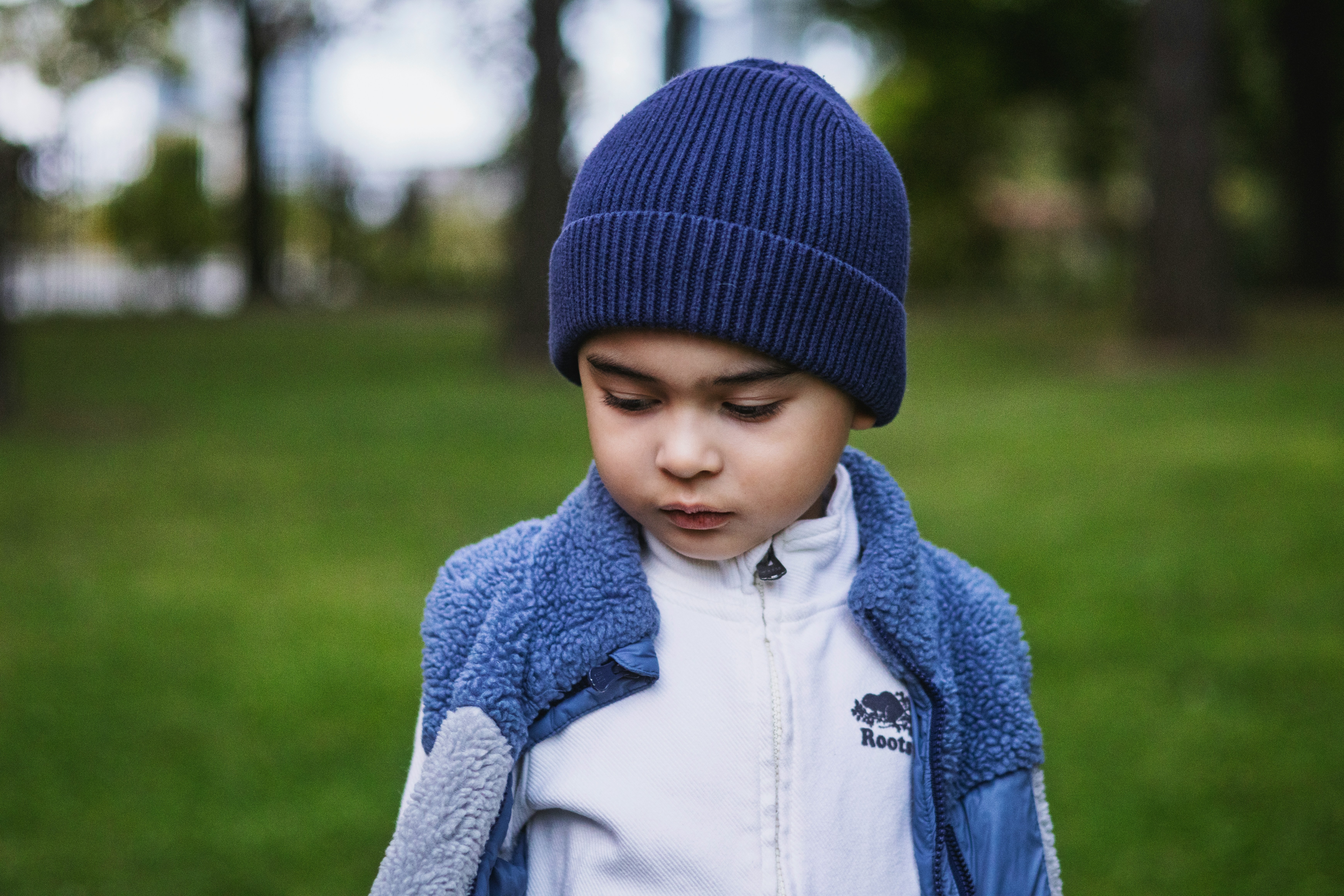 Menino de pele clara e cabelo escuro, usando gorro azul marinho, jaqueta azul com forro de lã e blusa branca com zíper, olhando para baixo em um parque com grama verde e árvores desfocadas ao fundo