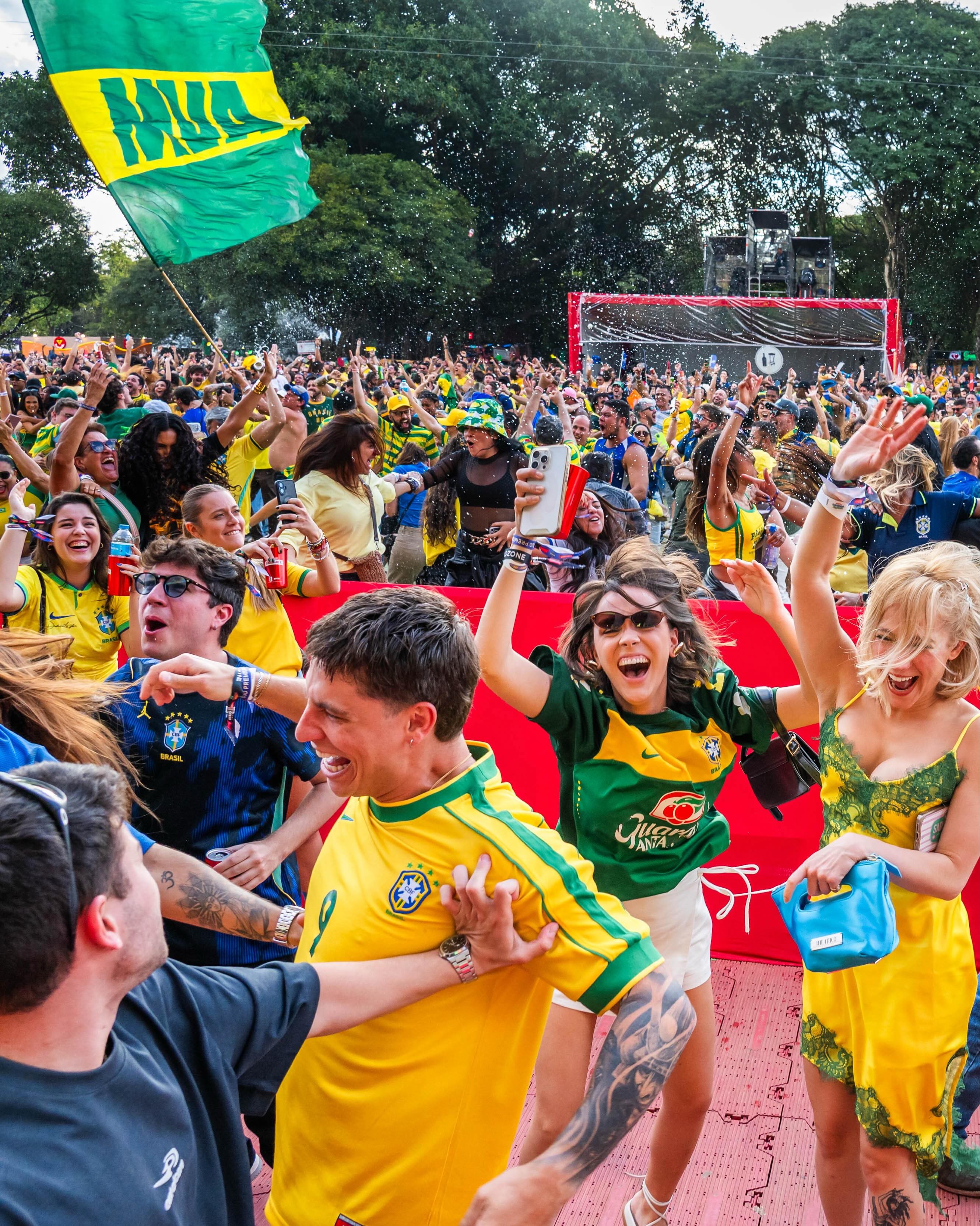Multidão de torcedores brasileiros, a maioria vestindo camisas amarelas e verdes, comemorando e levantando os braços em um evento ao ar livre. Um homem de camisa amarela e verde sorri enquanto é abraçado por outro. Duas mulheres ao lado dele, uma de camisa verde e outra de vestido amarelo, também sorriem e levantam os braços. Ao fundo, uma bandeira do Brasil tremula e um palco com equipamentos de som é visível.