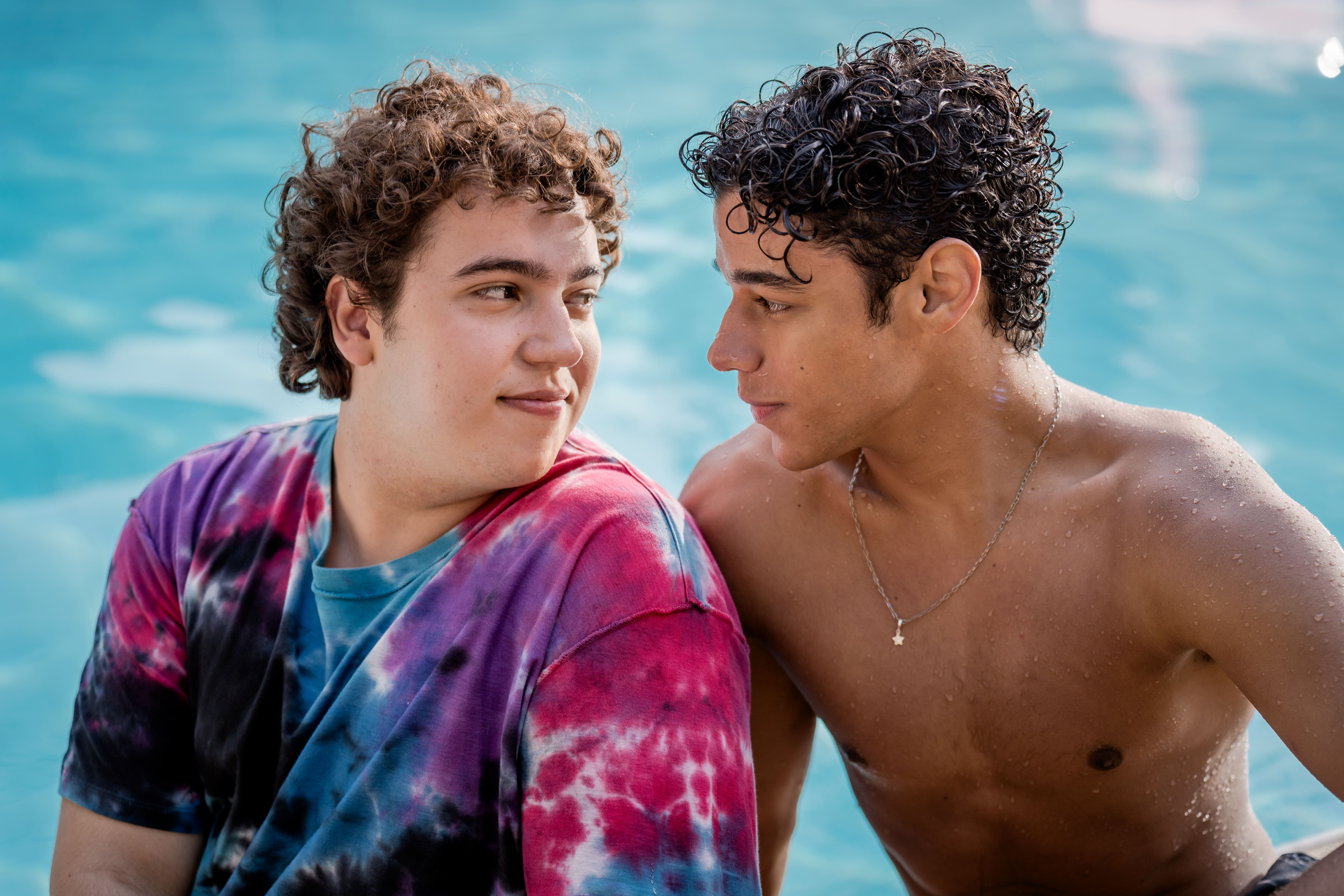 Dois jovens com cabelos cacheados, um de camiseta tie-dye e outro sem camisa, olham-se sorrindo em uma piscina