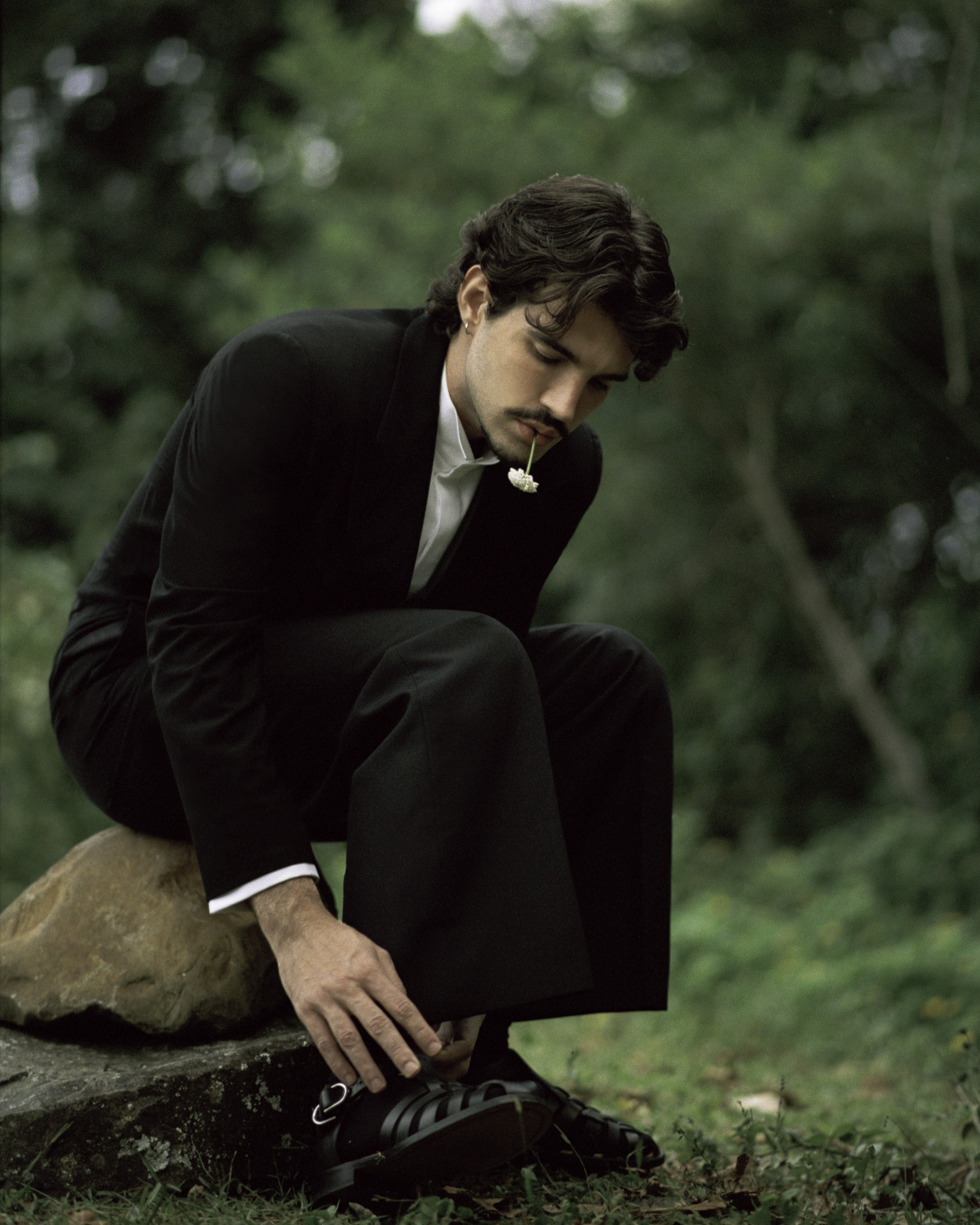 Homem de cabelo escuro e bigode, vestindo terno preto e camisa branca, agachado sobre uma rocha, ajustando a sandália preta. Ele tem uma pequena flor branca na boca, em um cenário de folhagem verde