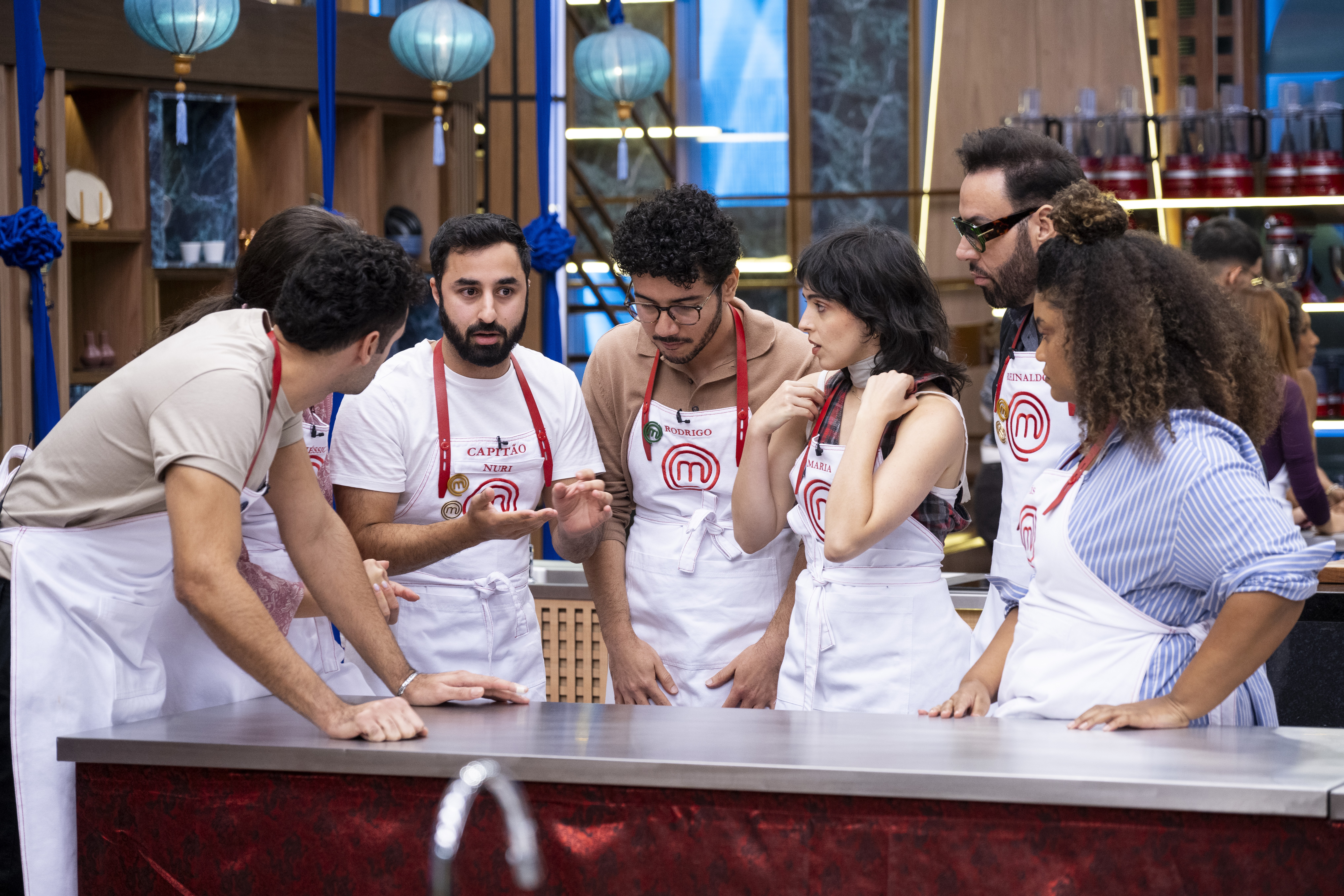 Grupo de sete pessoas, incluindo homens e mulheres, usando aventais brancos com detalhes vermelhos e o logo do MasterChef, reunidos em uma bancada de cozinha. Eles parecem estar em uma competição culinária, conversando e gesticulando, com um fundo de estantes e luminárias azuis