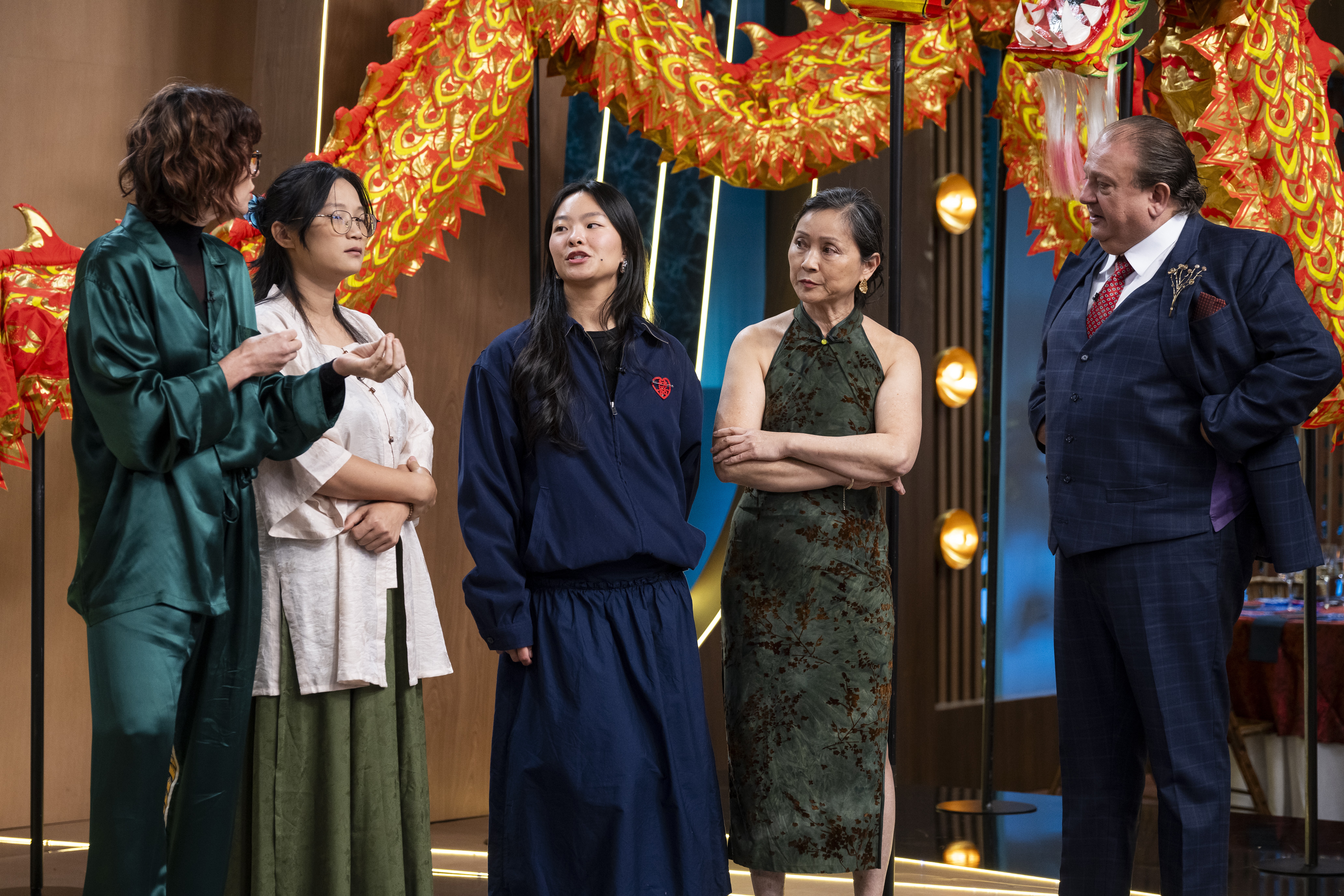 Cinco pessoas, três mulheres e dois homens, em um cenário com decorações de dragões chineses. À esquerda, uma mulher de cabelo cacheado e camisa verde-esmeralda conversa com uma mulher de óculos, camisa branca e saia verde-oliva. Ao centro, uma mulher de cabelo liso e conjunto azul-marinho olha para cima. À direita, uma mulher de vestido verde estampado e braços cruzados observa um homem de terno azul-marinho e gravata vermelha, que parece falar com ela