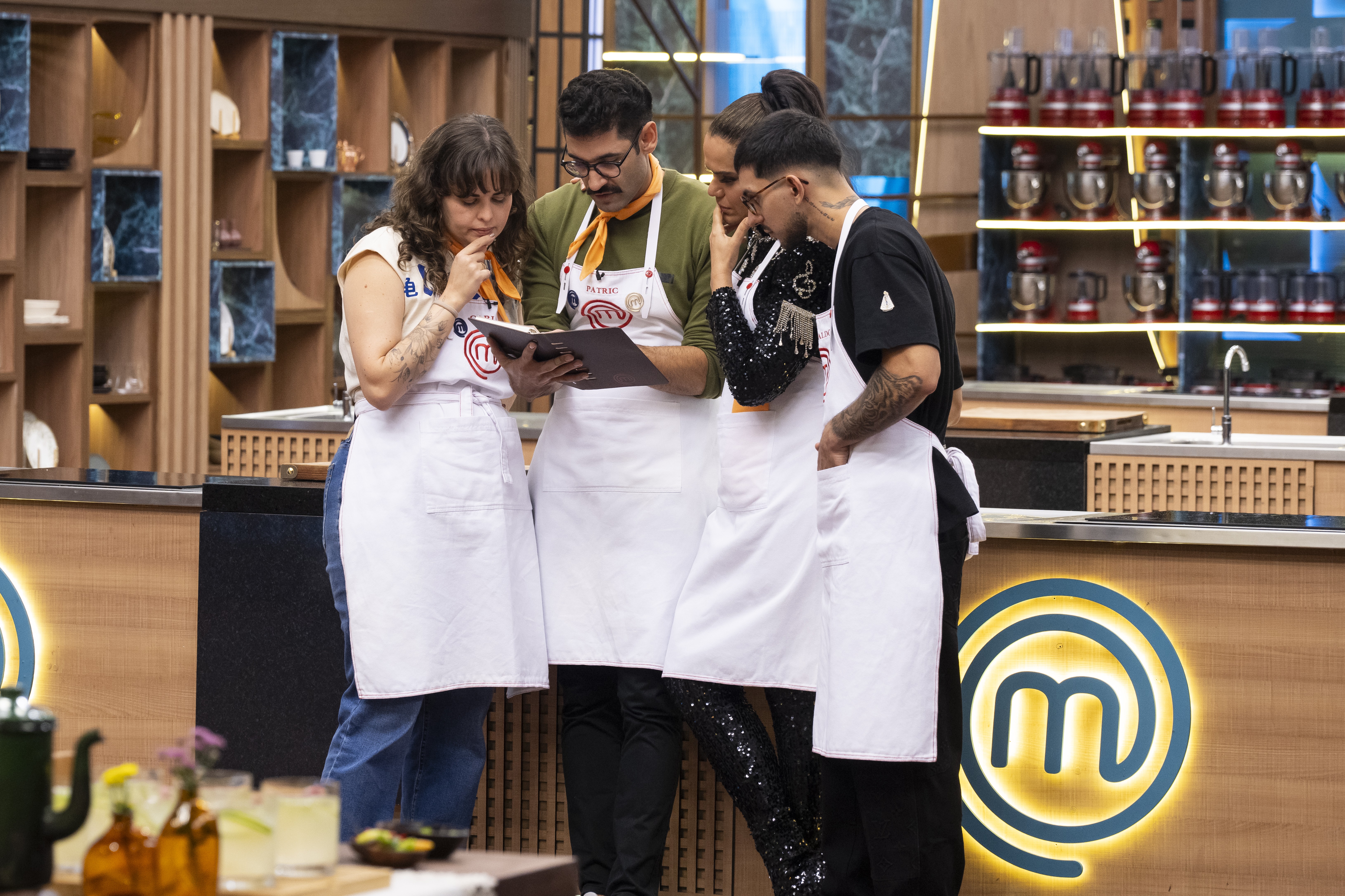 Quatro chefs, três homens e uma mulher, vestindo aventais brancos, olham atentamente para um caderno na cozinha do MasterChef. A mulher à esquerda tem a mão no queixo, pensativa. O homem central, de bigode e óculos, segura o caderno. Ao fundo, prateleiras de madeira com objetos decorativos e utensílios de cozinha. O logo do MasterChef brilha na bancada.