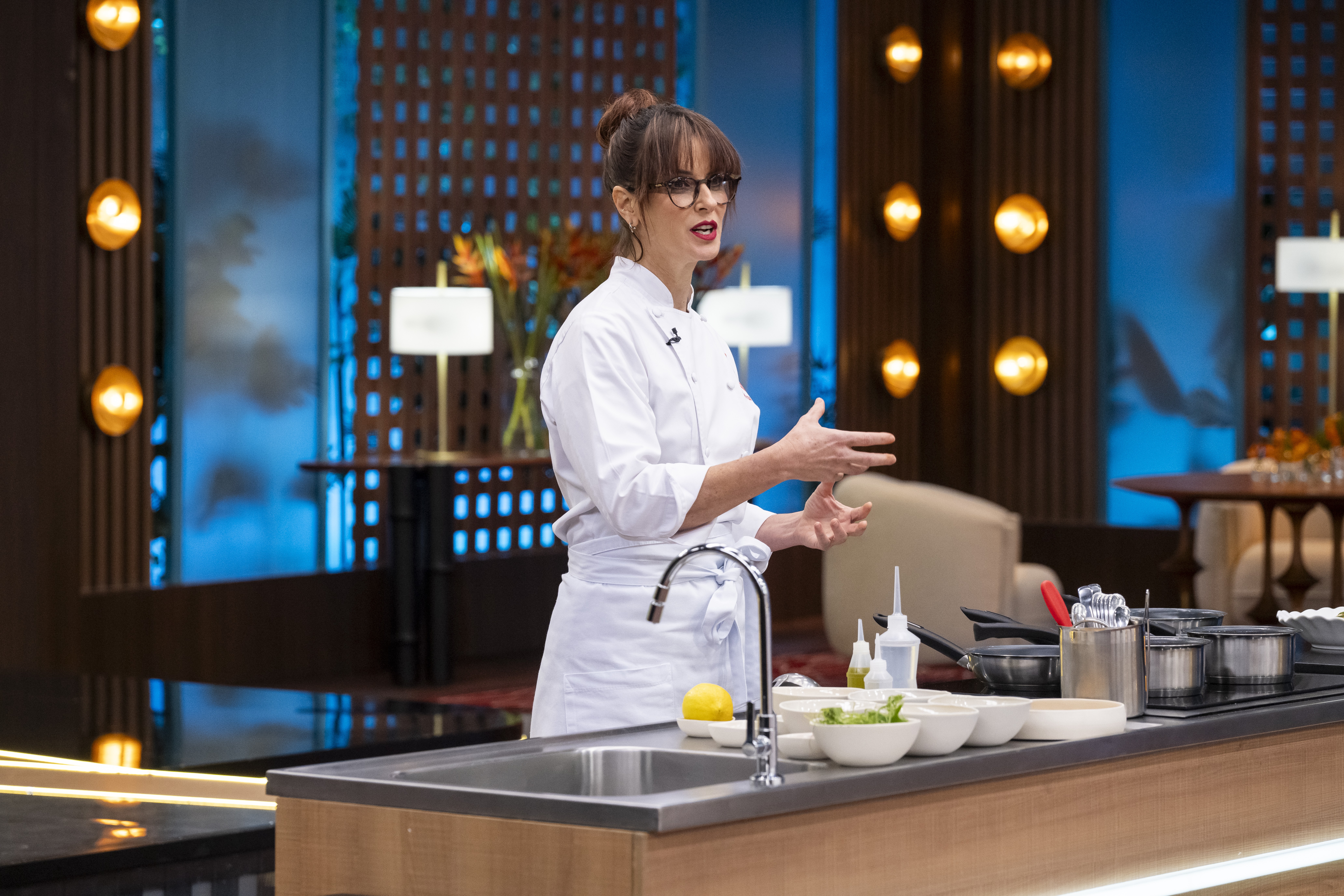 Mulher de óculos, cabelo preso e uniforme de chef branco, gesticula com as mãos abertas em um estúdio de cozinha. Ela está atrás de uma bancada com pia, limão, tigelas e panelas. Ao fundo, painéis de madeira com luzes douradas e um ambiente com mesas e cadeiras.