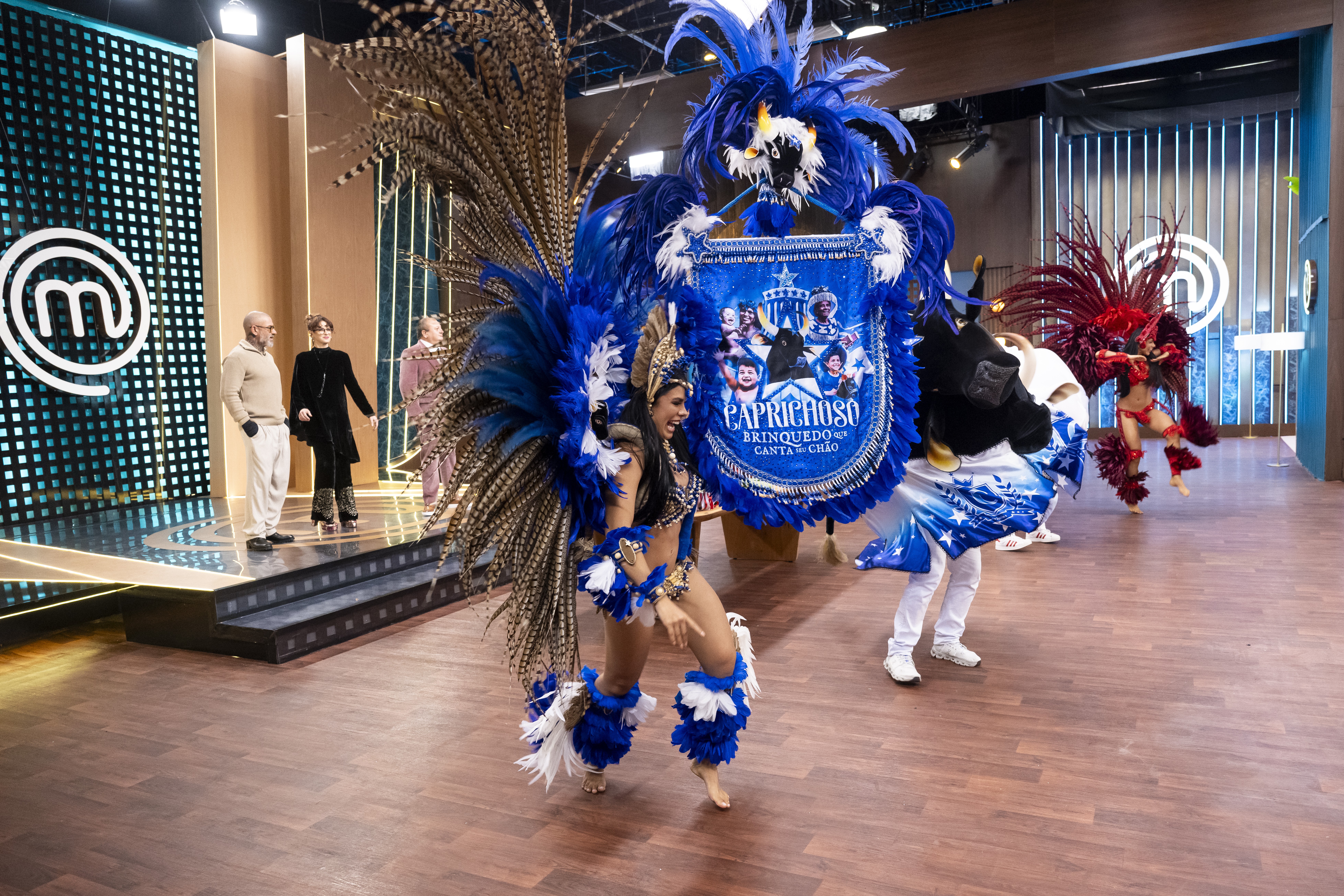 Mulher sorridente em traje de carnaval azul e marrom com plumas, dançando no centro de um palco. Ao fundo, três pessoas assistem, e à direita, outra dançarina de vermelho.