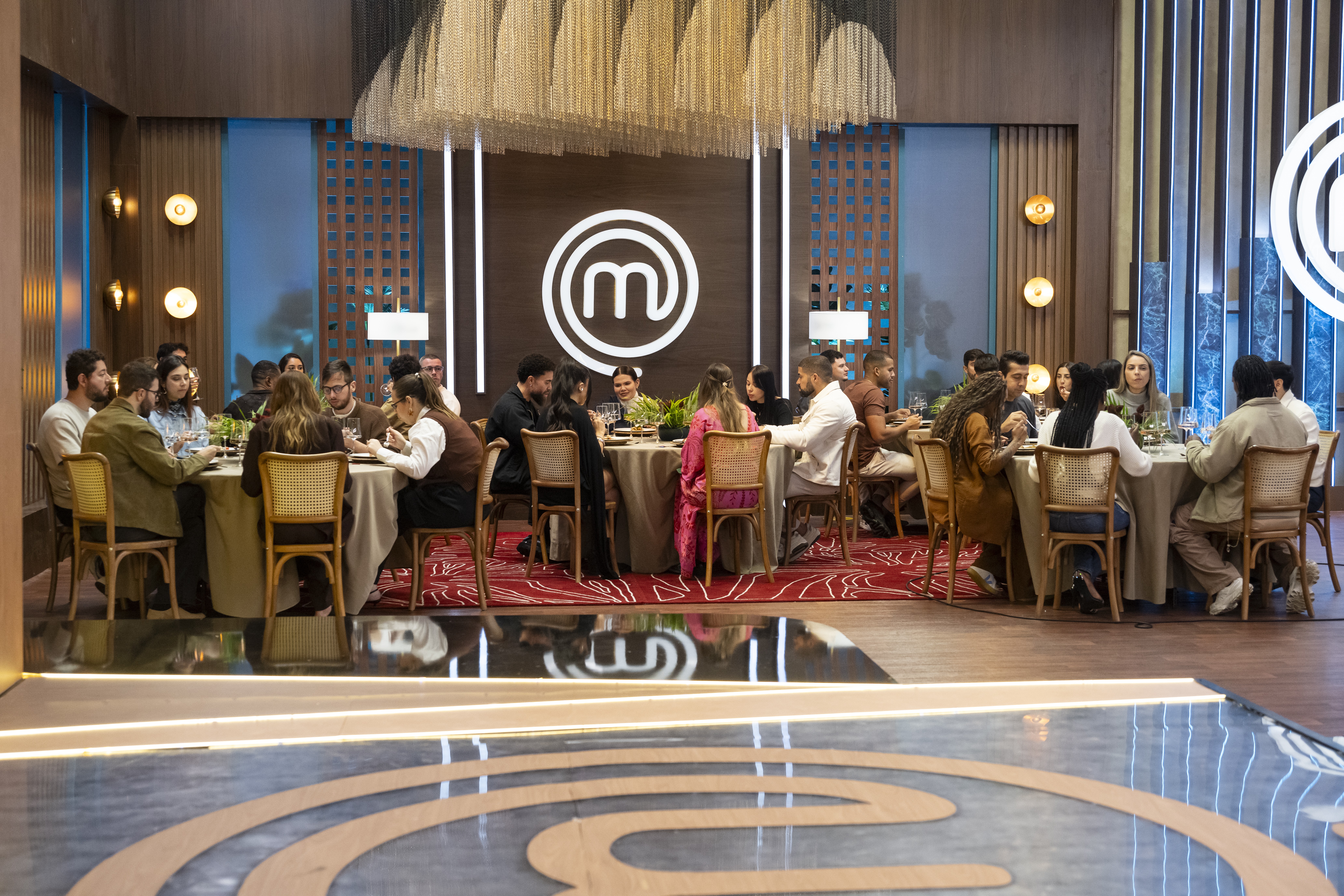 Pessoas sentadas em mesas redondas, comendo e conversando, em um ambiente de restaurante com iluminação aconchegante e o logo do MasterChef ao fundo
