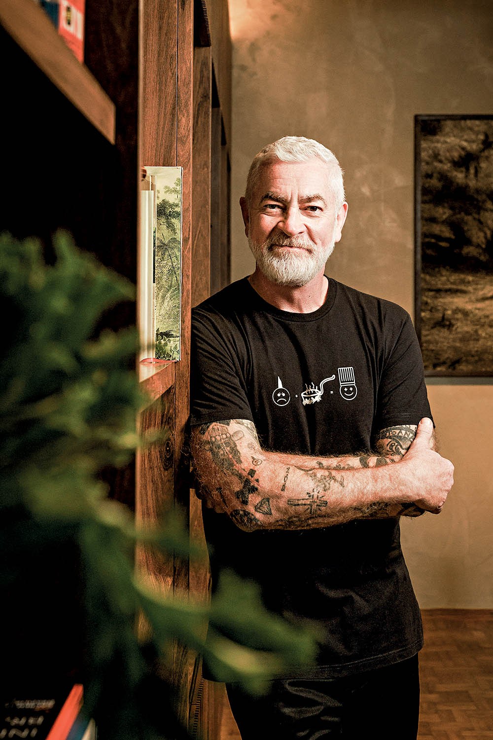 Homem branco de barba e cabelo grisalhos, sorrindo, com braços tatuados cruzados, vestindo camiseta preta com estampas de emojis e um chapéu de chef, encostado em uma estante de madeira com livros e plantas