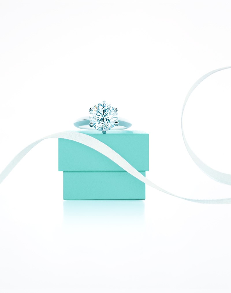 Anel de diamante solitário com lapidação brilhante em aro de prata, sobre uma caixa turquesa e fita branca, em fundo branco