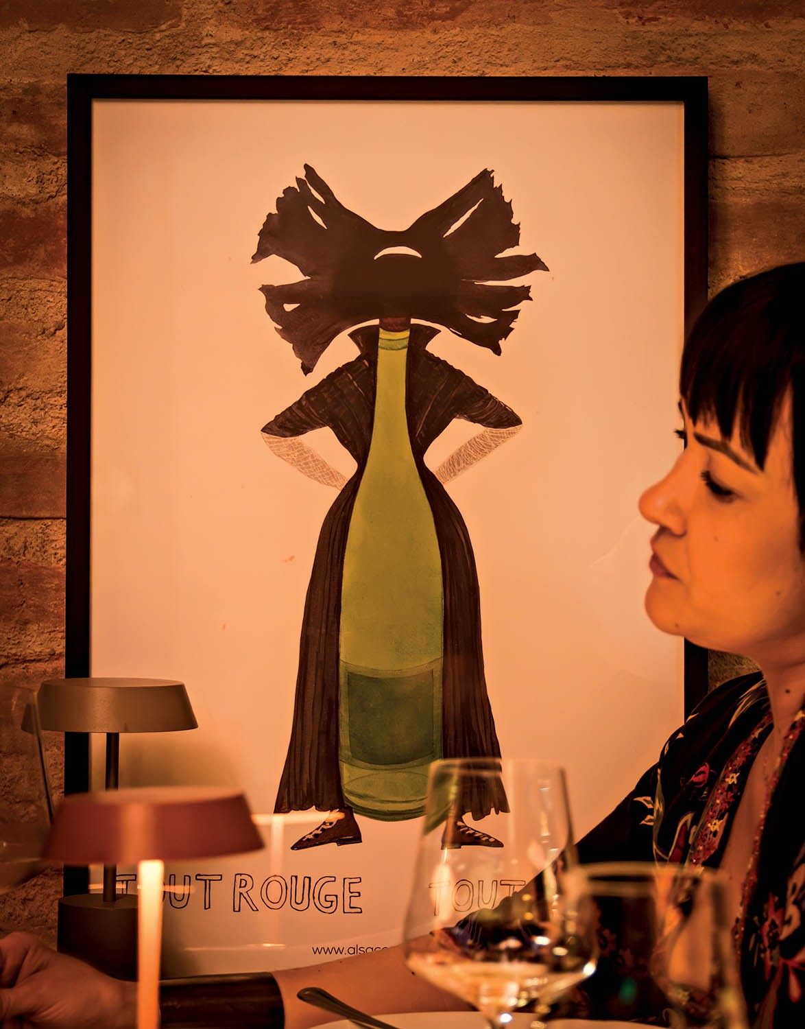 Mulher de perfil, com cabelo escuro e franja, sentada à mesa com taças de vinho e uma luminária. Ao fundo, um pôster com uma garrafa de vinho verde vestindo um casaco escuro e sapatos, com uma cabeça abstrata preta e texto CHUT ROUGE e www.alsace.com