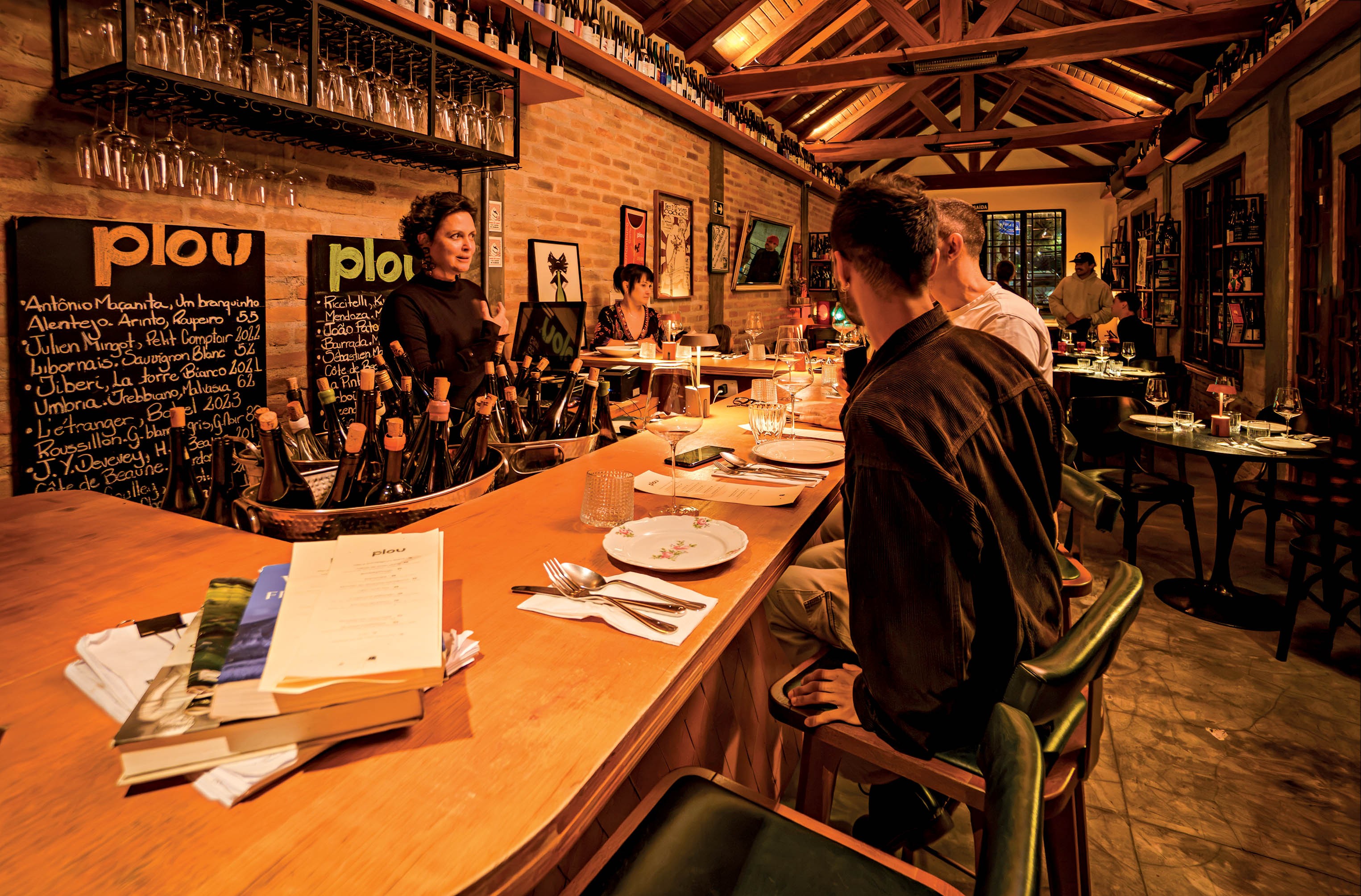 Mulher de cabelo cacheado escuro e blusa preta atende clientes em um bar de madeira com garrafas de vinho e quadros de giz com menus. Clientes sentados em bancos altos e mesas ao fundo, sob iluminação quente e teto de madeira.