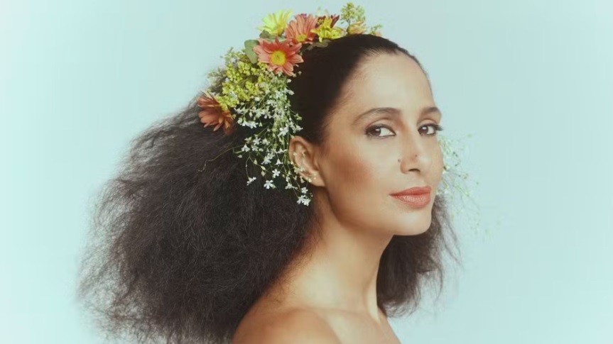 Mulher de pele morena, cabelo crespo escuro e solto, com uma coroa de flores coloridas na cabeça, olhando para a frente com expressão serena, ombro nu, fundo azul claro