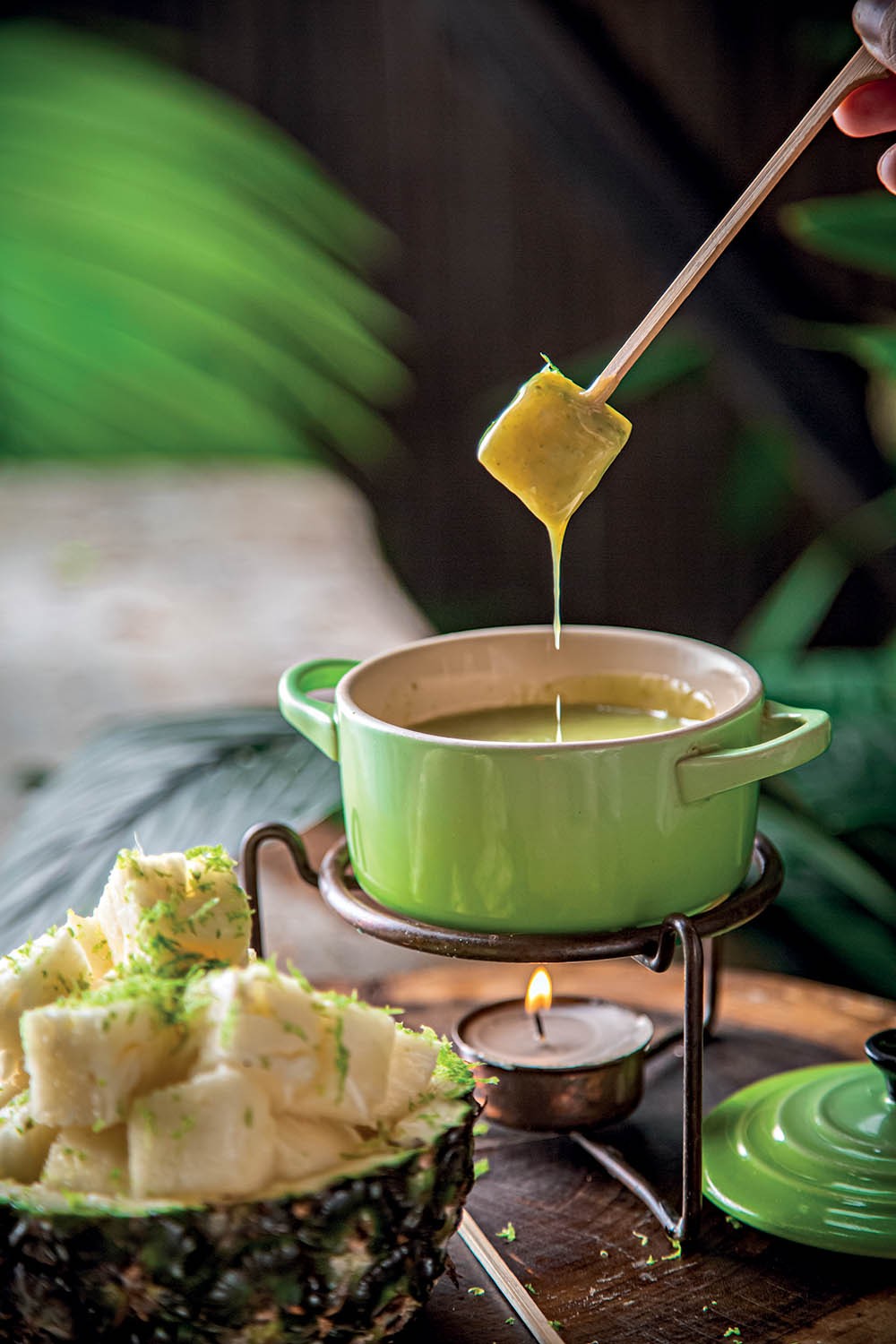 Uma mão segura um espeto de bambu com um pedaço de abacaxi mergulhado em um fondue verde claro, que escorre da fruta. O fondue está em uma panela verde sobre um rechaud com uma vela acesa. Ao lado, uma metade de abacaxi com pedaços da fruta picados e raspas de limão. Folhas verdes desfocadas ao fundo