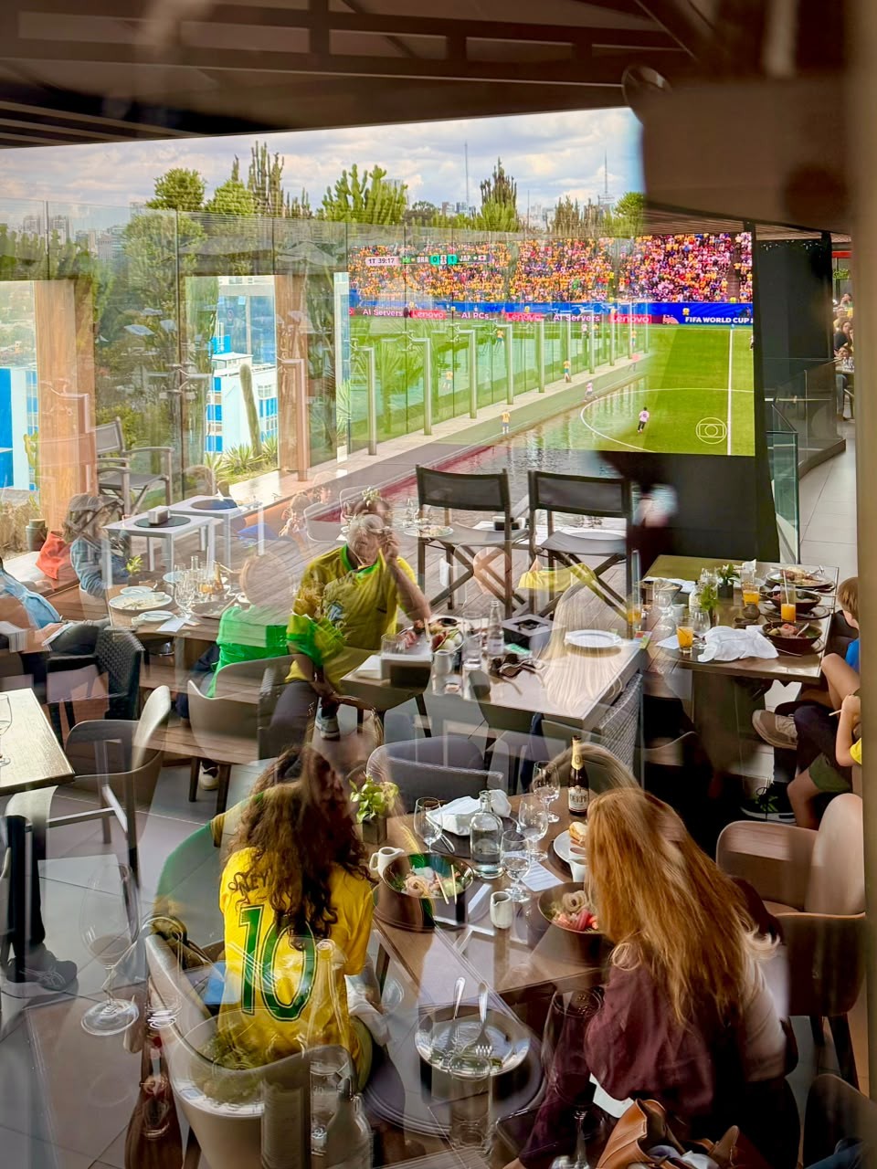 Pessoas assistem a um jogo de futebol em um telão gigante, refletido em uma janela de um restaurante. Uma mulher de camisa amarela com o número 10 está sentada de costas, enquanto outra pessoa de camisa amarela e verde observa o telão. Mesas com pratos e bebidas estão dispostas no ambiente.